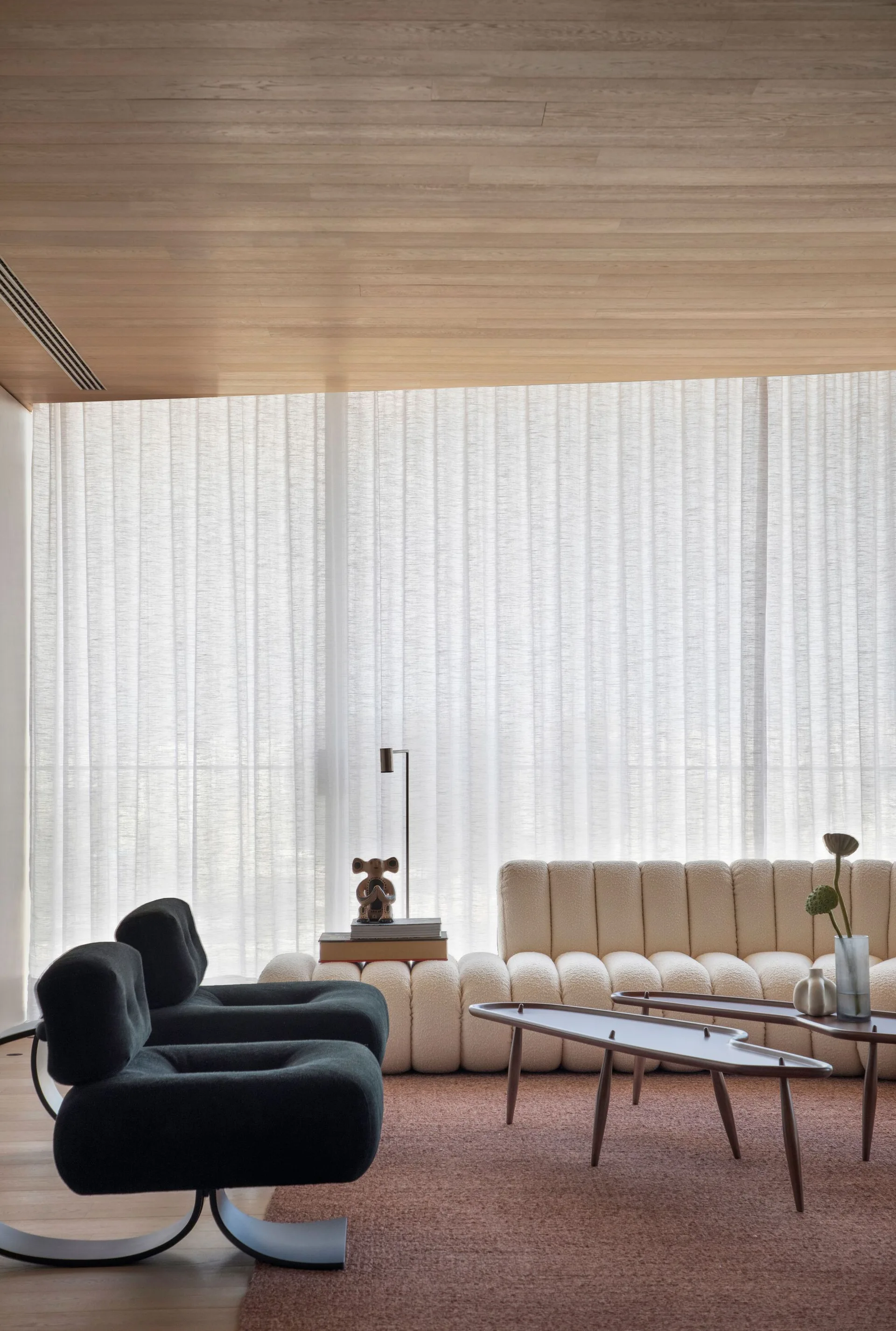 A living room features a low off-white channel-tufted sofa, two dark grey armchairs, and organic wood tables set against sheer white curtains.