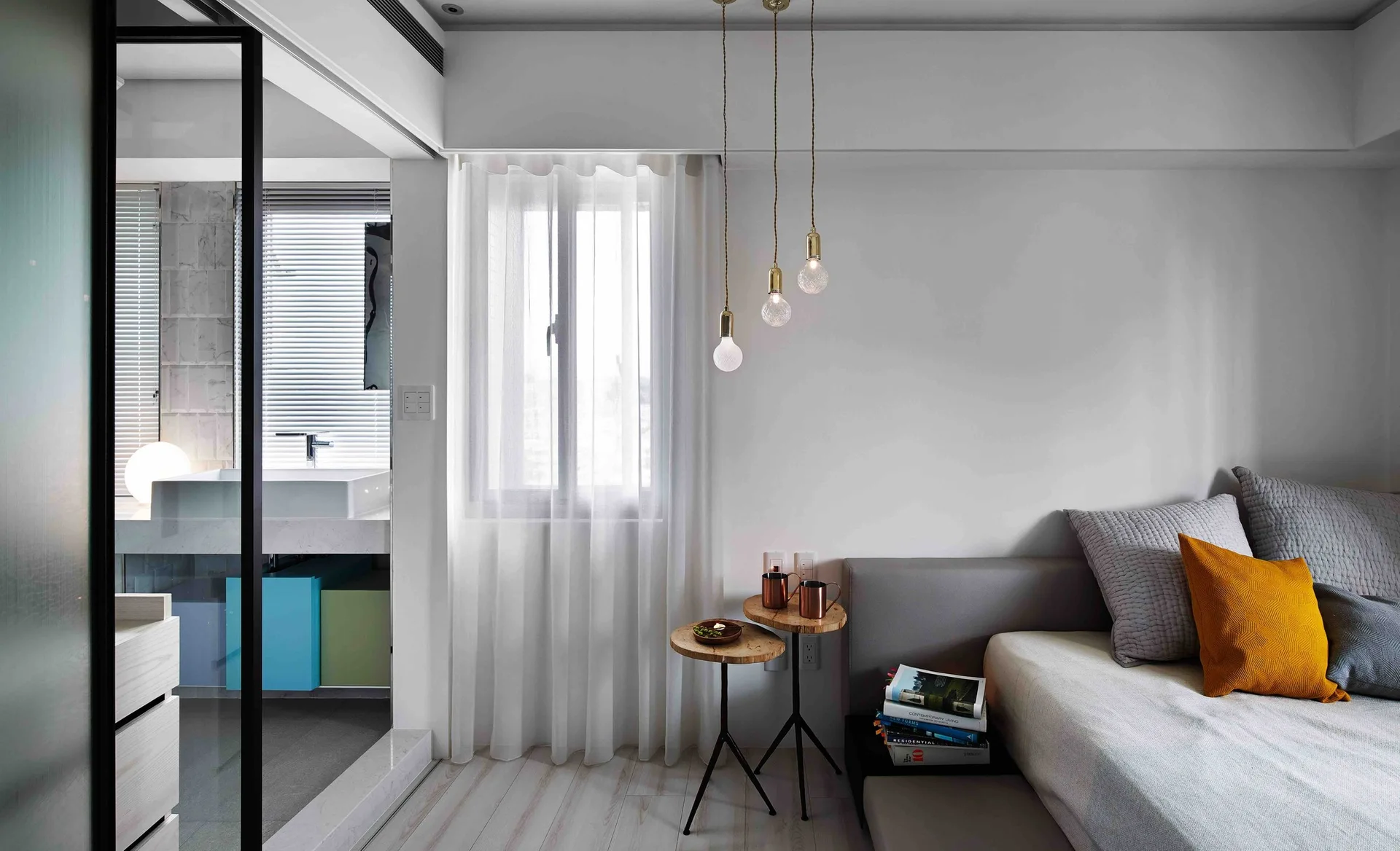 A minimalist bedroom featuring a platform bed with gray and mustard pillows, sheer white curtains, and a black-framed glass partition to the bathroom.