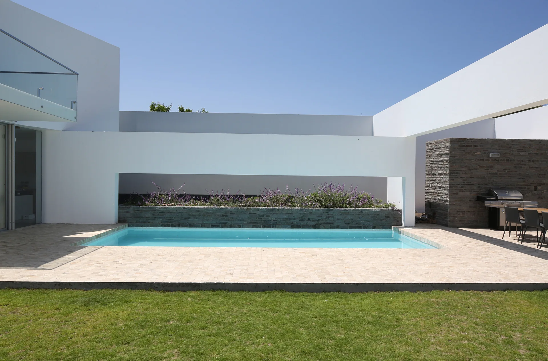 A rectangular swimming pool set in a tiled beige patio is framed by modern white geometric structures, featuring a raised stone planter with purple flowers.