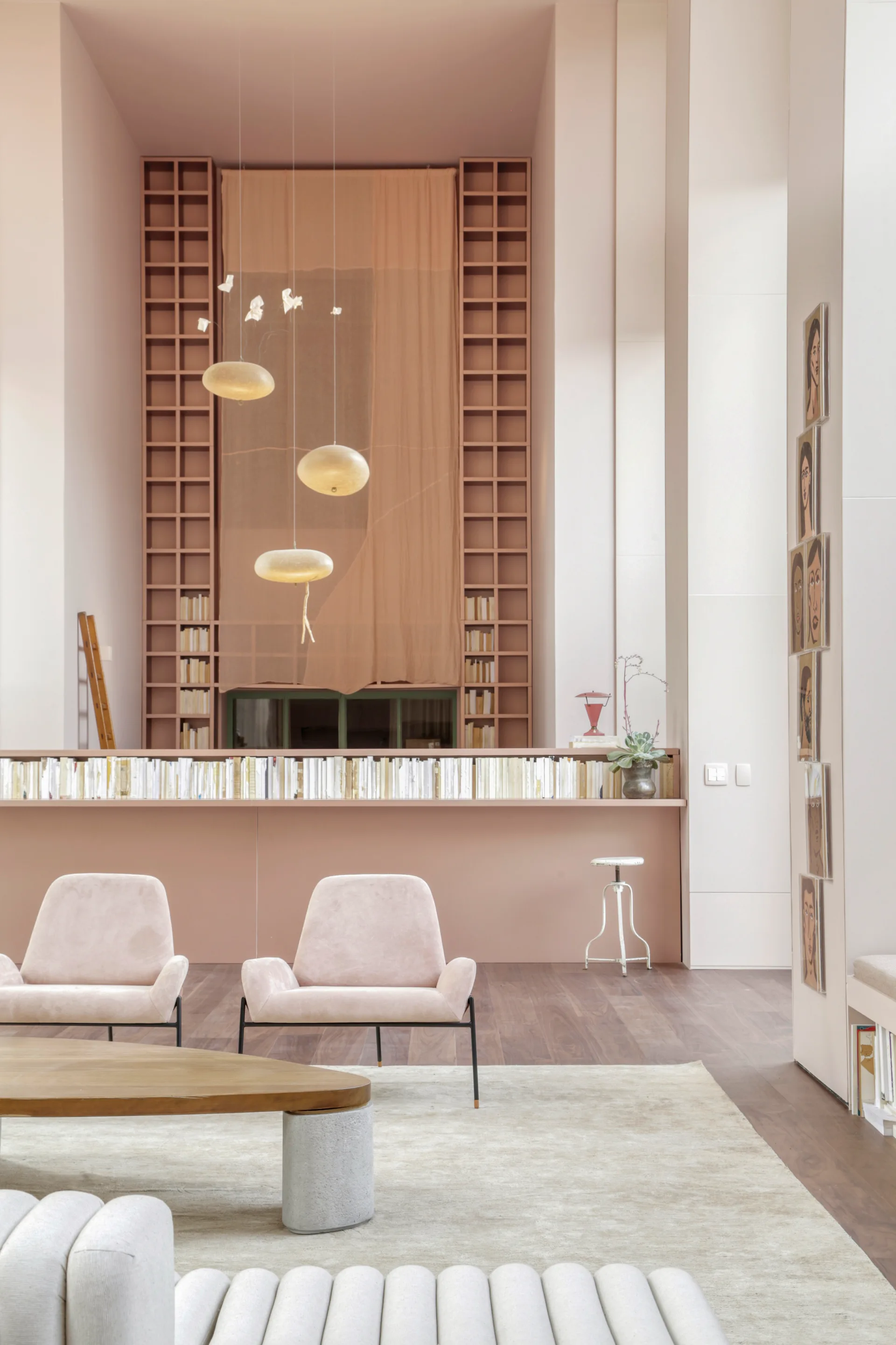 A high-ceilinged living room features blush pink walls and built-in shelving, two low armchairs, and glowing sculptural pendant lights hanging overhead.