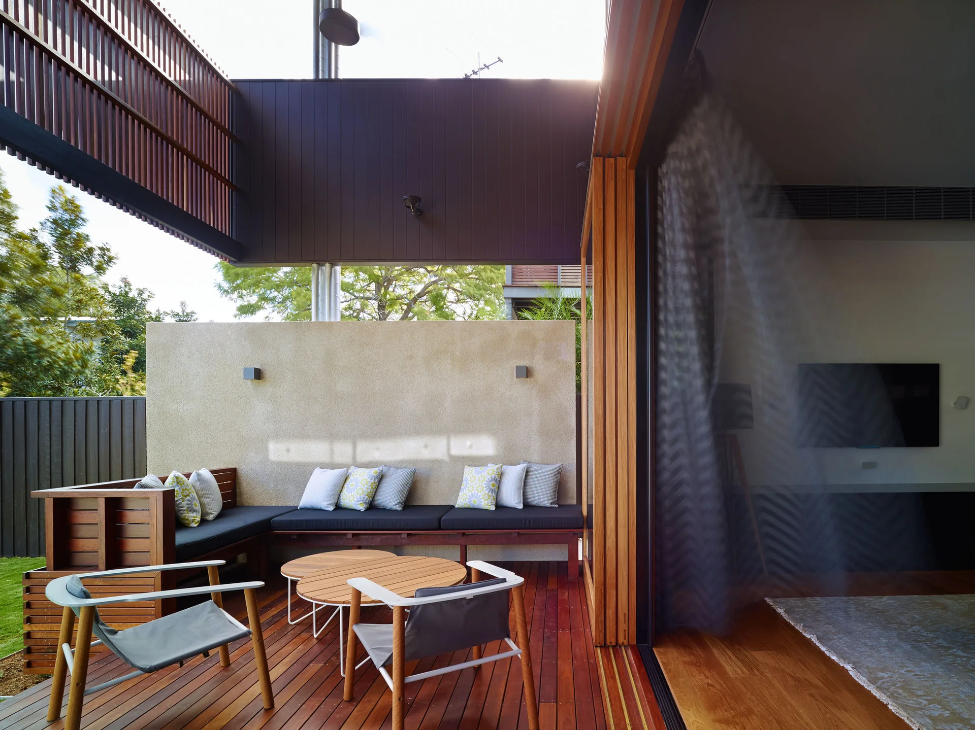 A modern wood deck features a built-in L-shaped bench with throw pillows, a textured stucco privacy wall, and sliding doors leading indoors.