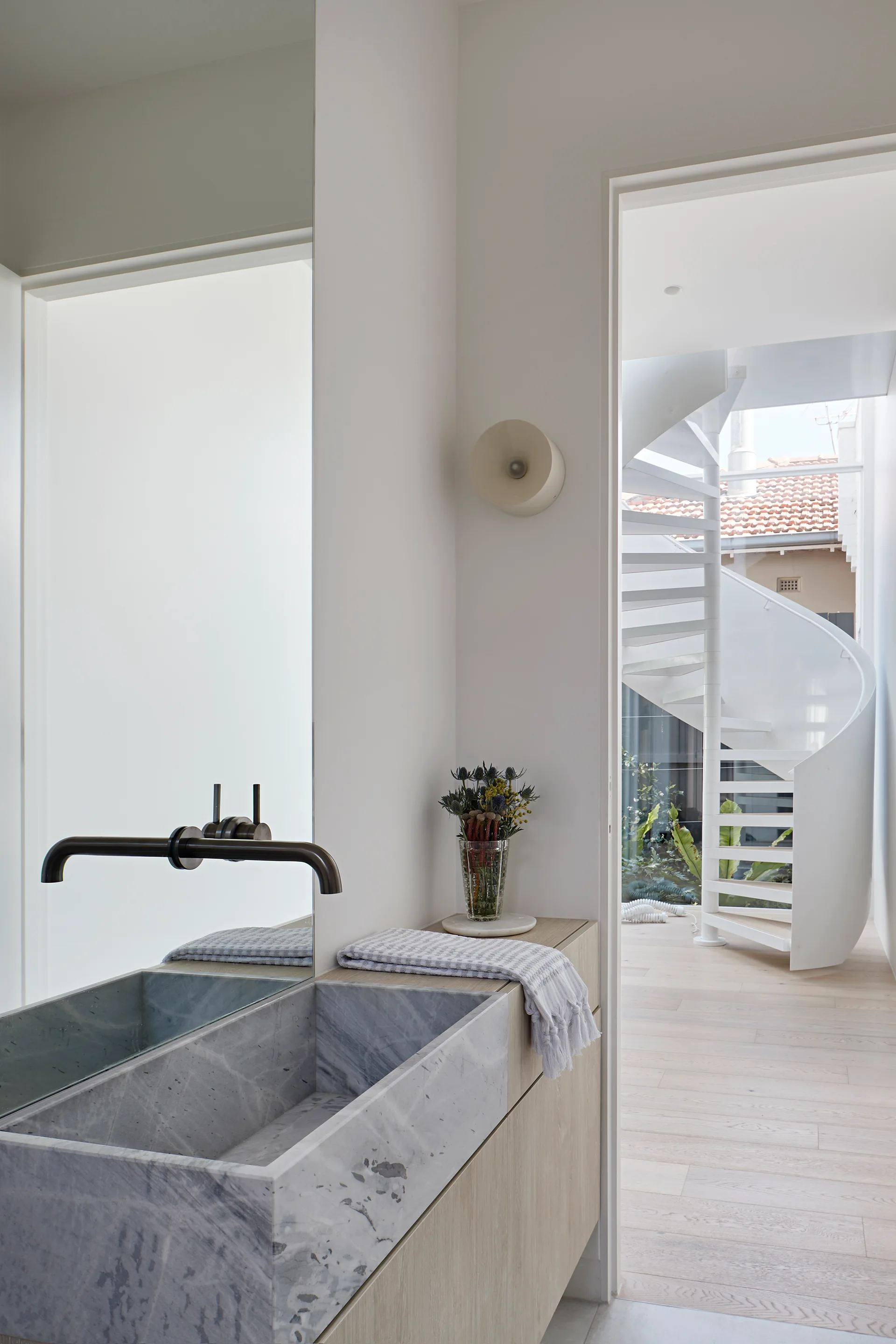 A modern bathroom features a grey veined marble trough sink, a light wood vanity, and a view through a doorway to a white spiral staircase.