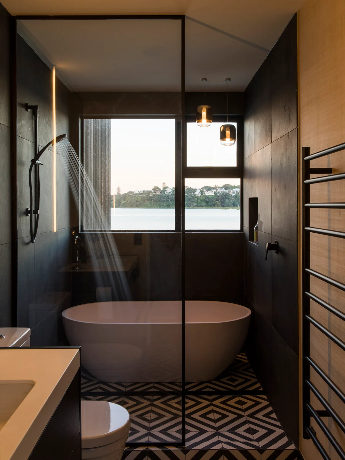 A contemporary bathroom featuring a white freestanding bathtub, black tiled walls, a glass shower partition, and black and white geometric floor tiles.