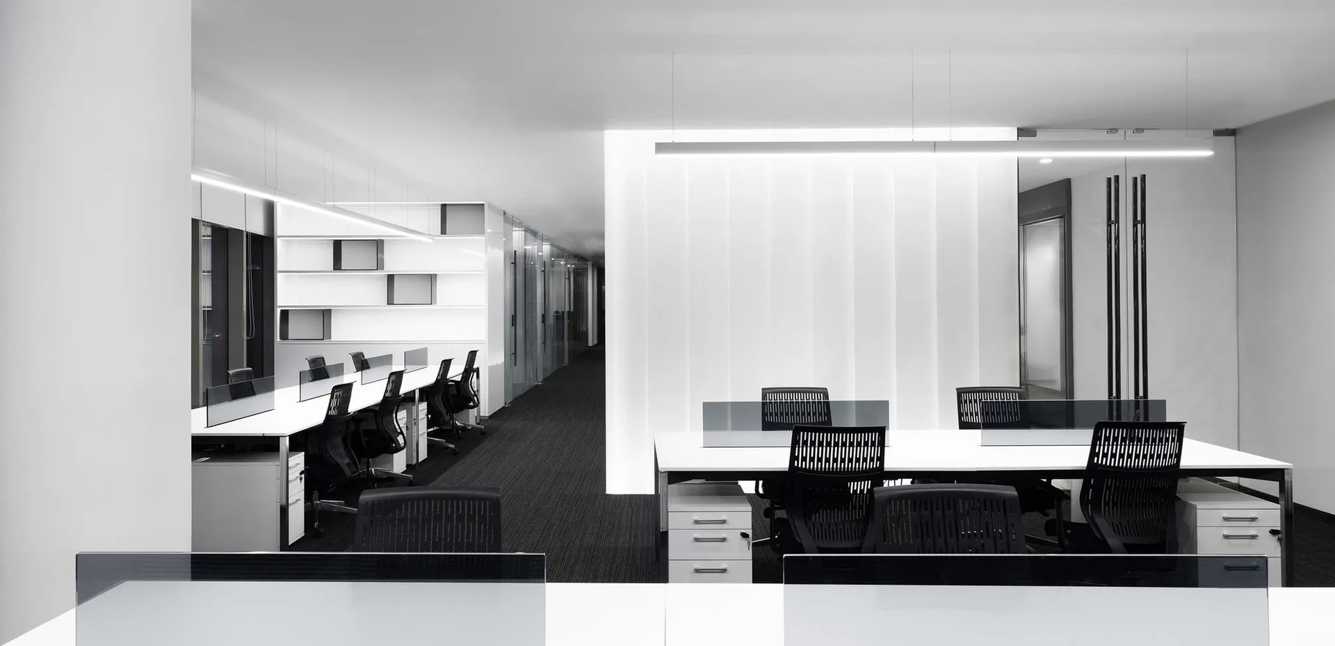 A minimalist office interior with white desks, black chairs, dark carpet, and linear overhead pendant lighting.