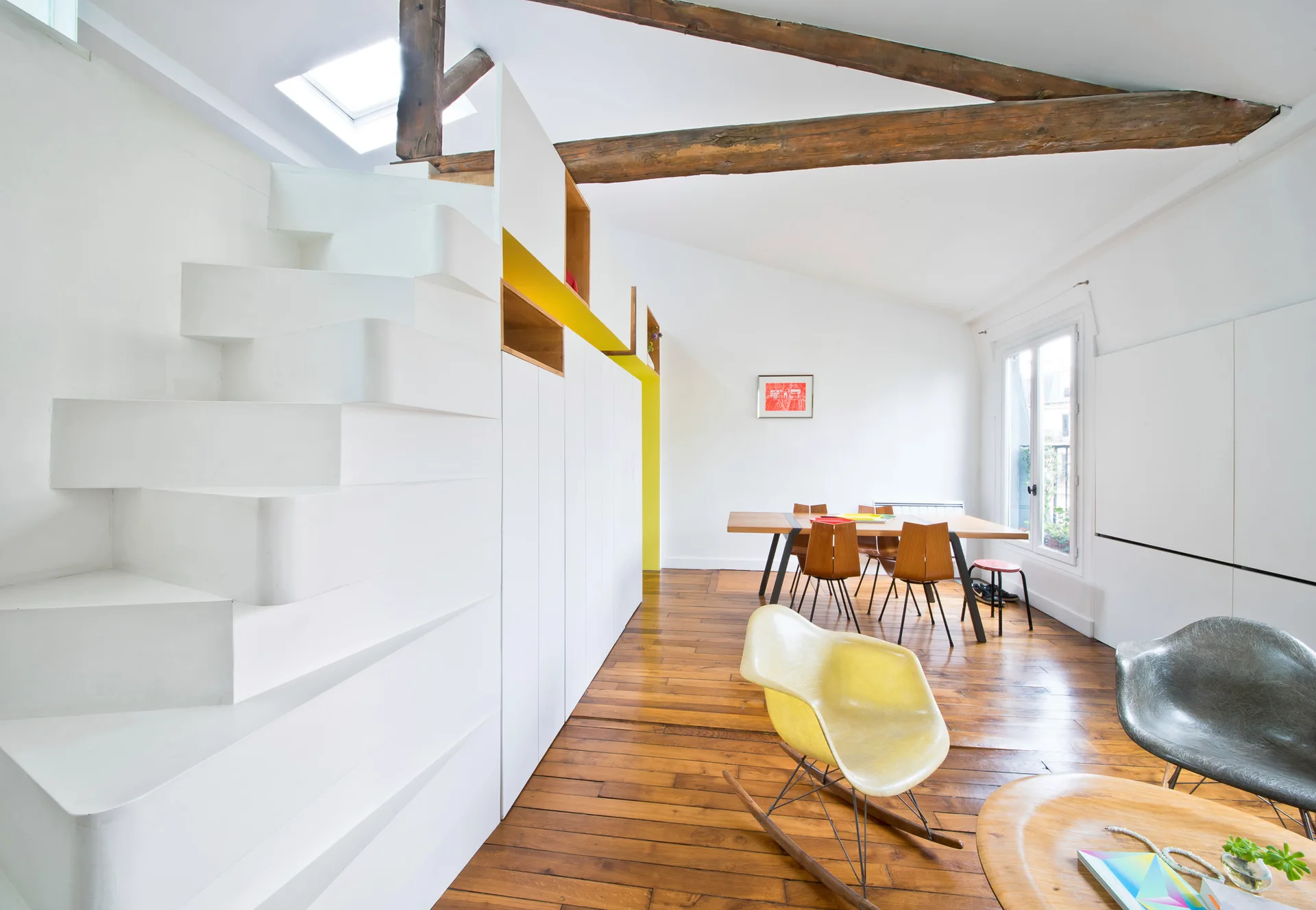 An open loft space features a sculptural white staircase integrated with wall storage, rustic ceiling beams, and a mid-century modern seating area.