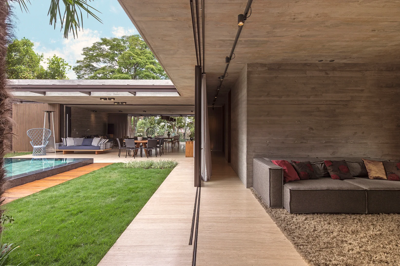 A modernist living space with board-formed concrete walls transitions to an outdoor courtyard with a grass lawn and swimming pool.