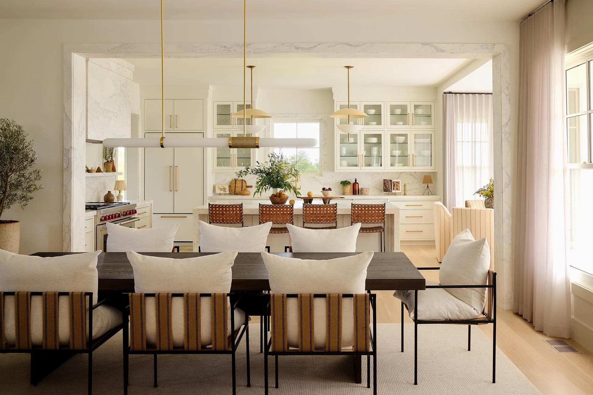 A dark wood dining table with woven strap chairs sits before a white kitchen framed by a marble-trimmed archway.