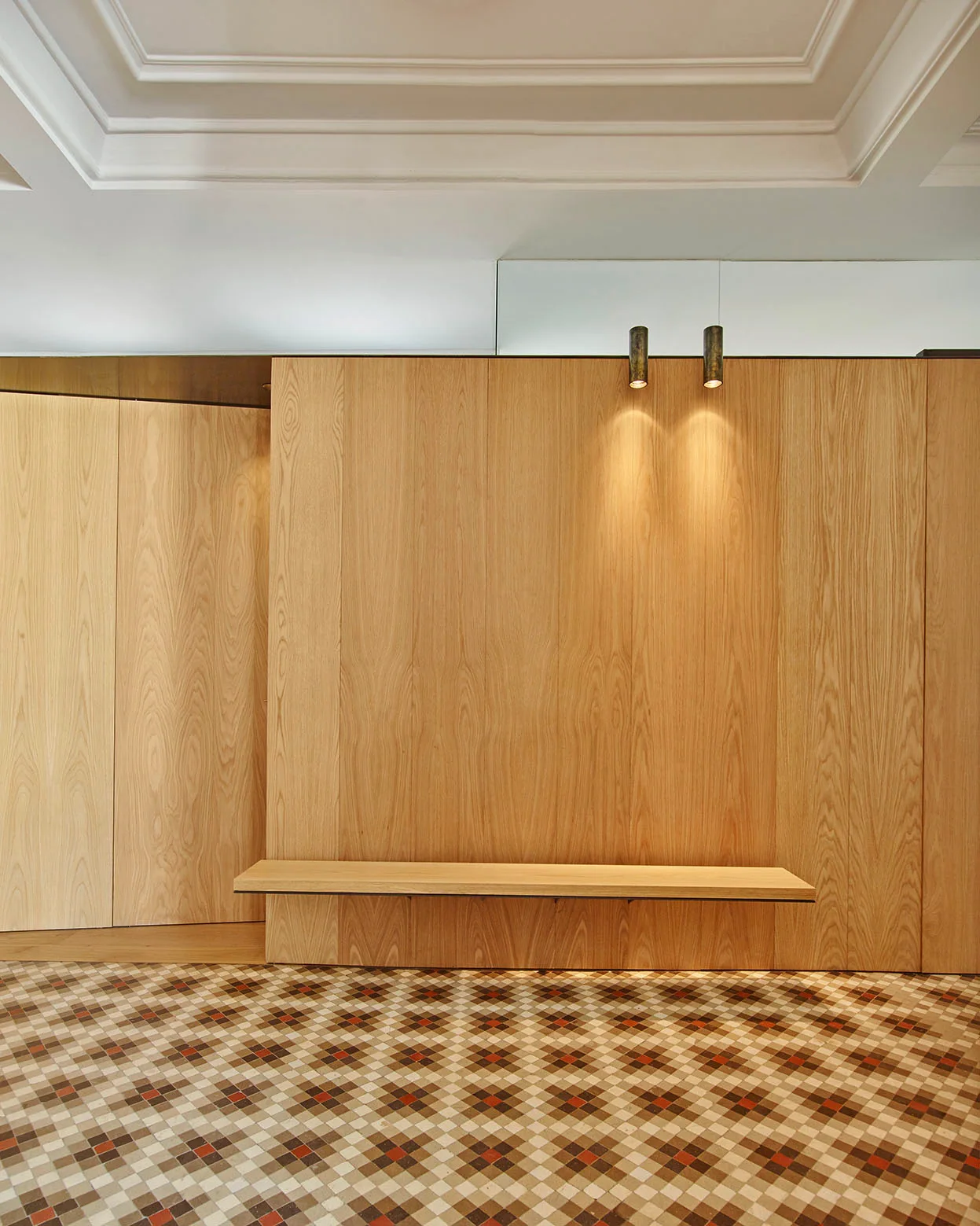 A minimalist entryway featuring a full wood-paneled wall, a matching floating wooden bench, and a geometric tiled floor beneath a stepped ceiling.
