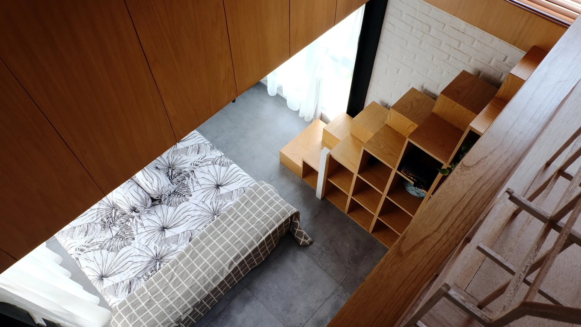 A high-angle view of a loft bedroom with a botanical print bed, grey tile floor, white brick wall, and a wooden storage staircase.