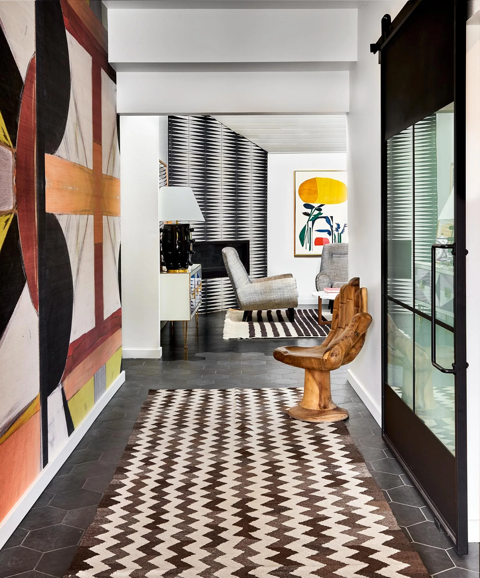 A modern entryway featuring a bold geometric wall mural, a chevron runner rug over dark tile, and a unique hand-shaped wooden chair.