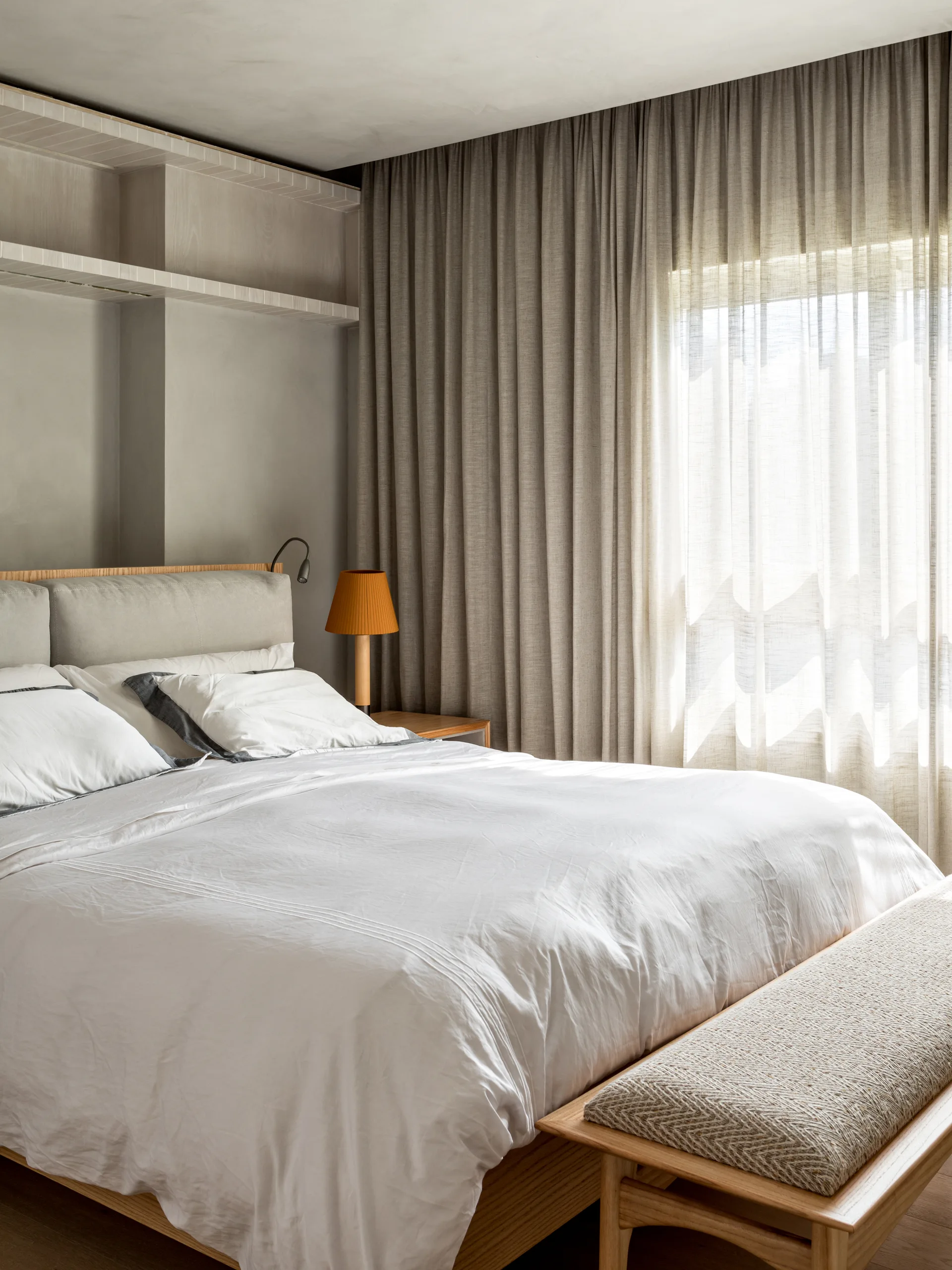 A minimalist bedroom featuring a wooden bed with gray upholstered headboard, a wooden bench at the foot, built-in wall shelving, and layered curtains.