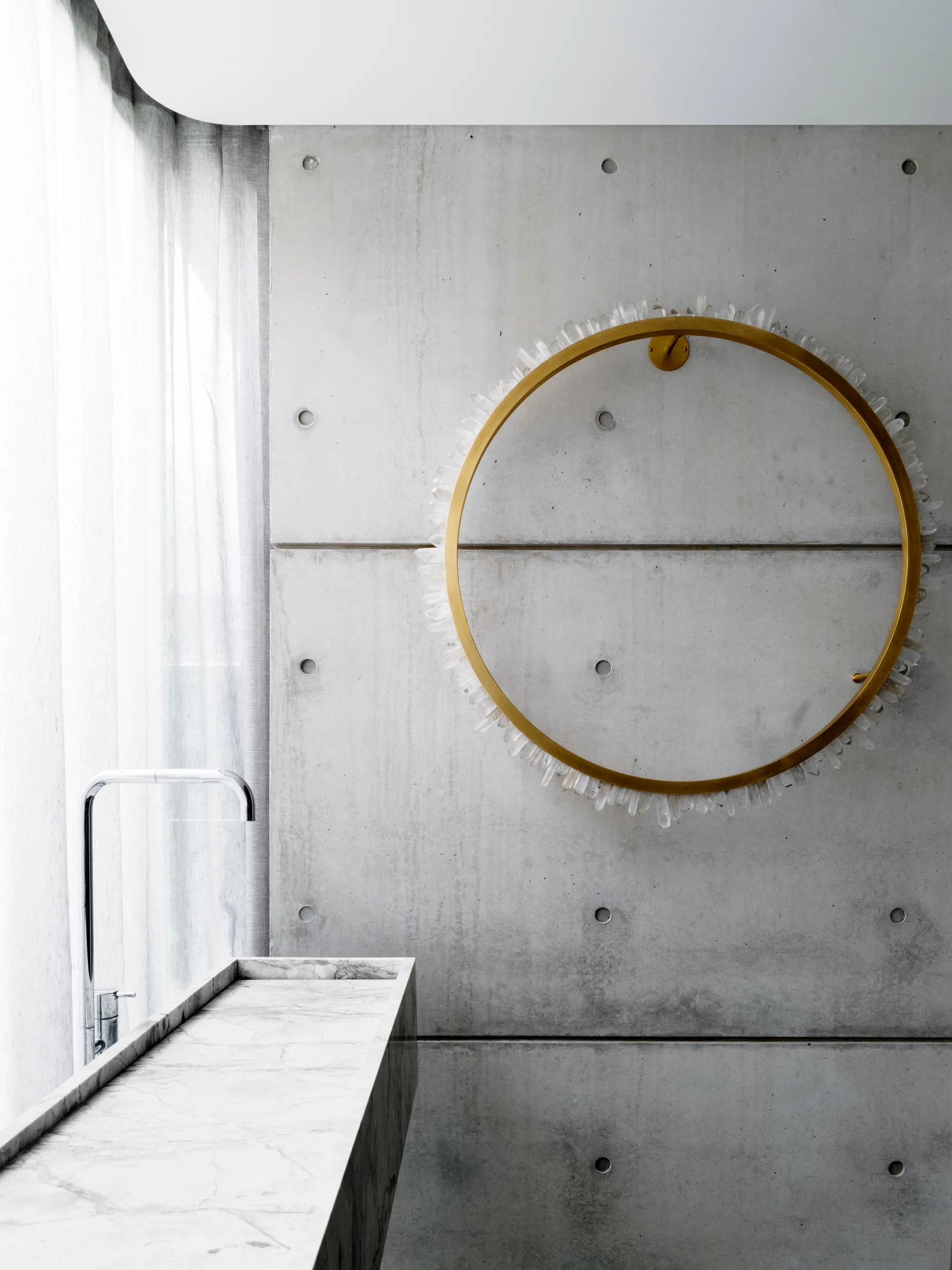 A marble vanity and chrome faucet stand before a raw concrete wall adorned with a circular gold mirror trimmed in quartz crystals.