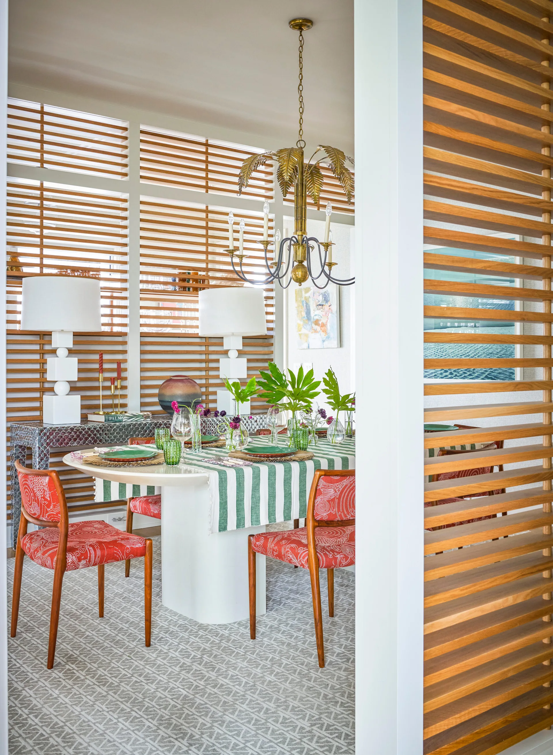 An eclectic dining room with red patterned chairs, a white pedestal table, a brass palm tree chandelier, and horizontal wood slat privacy screens.