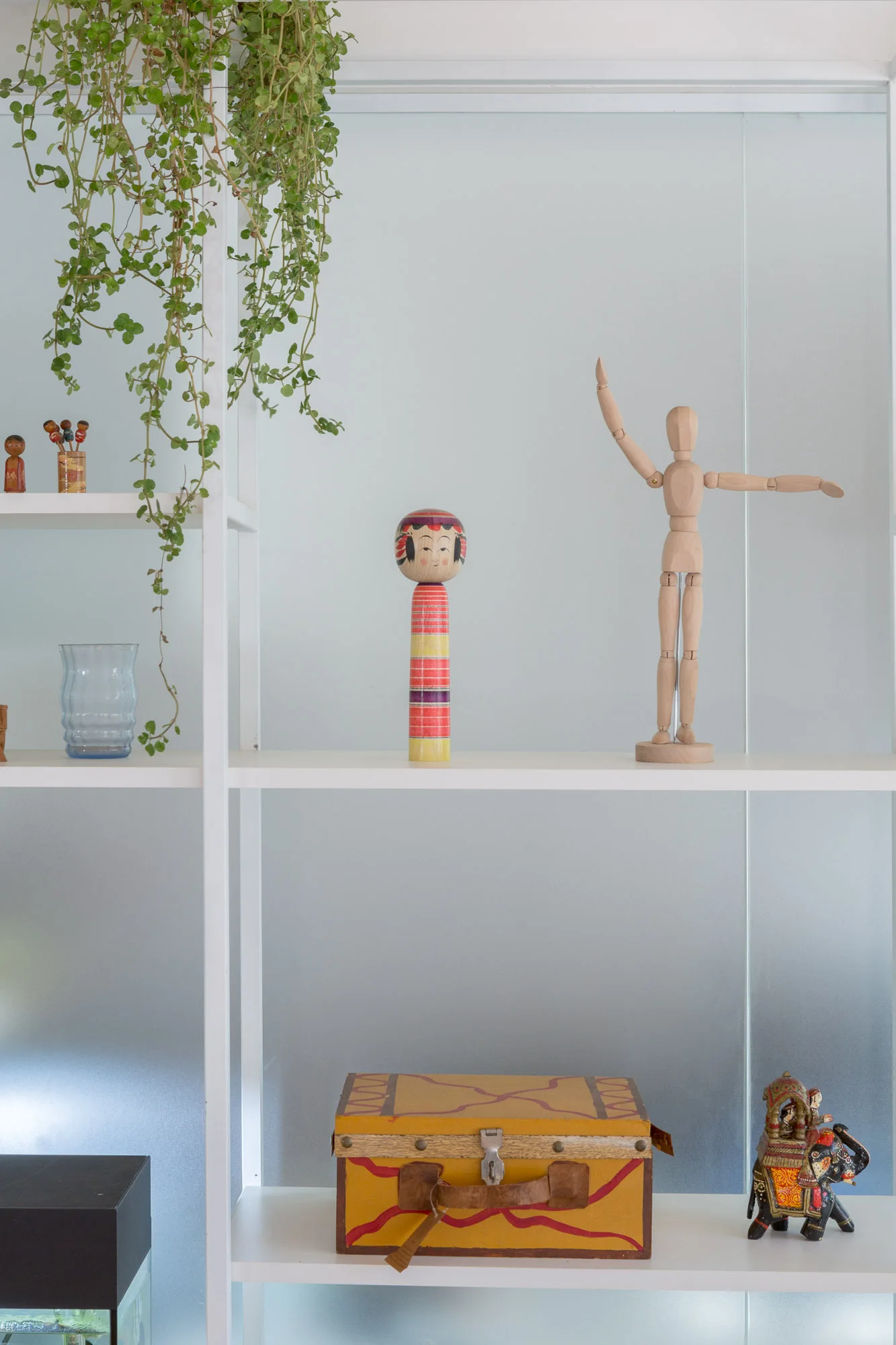 White open shelves display a trailing plant, a wooden mannequin, a kokeshi doll, and a yellow patterned box against a frosted glass wall.
