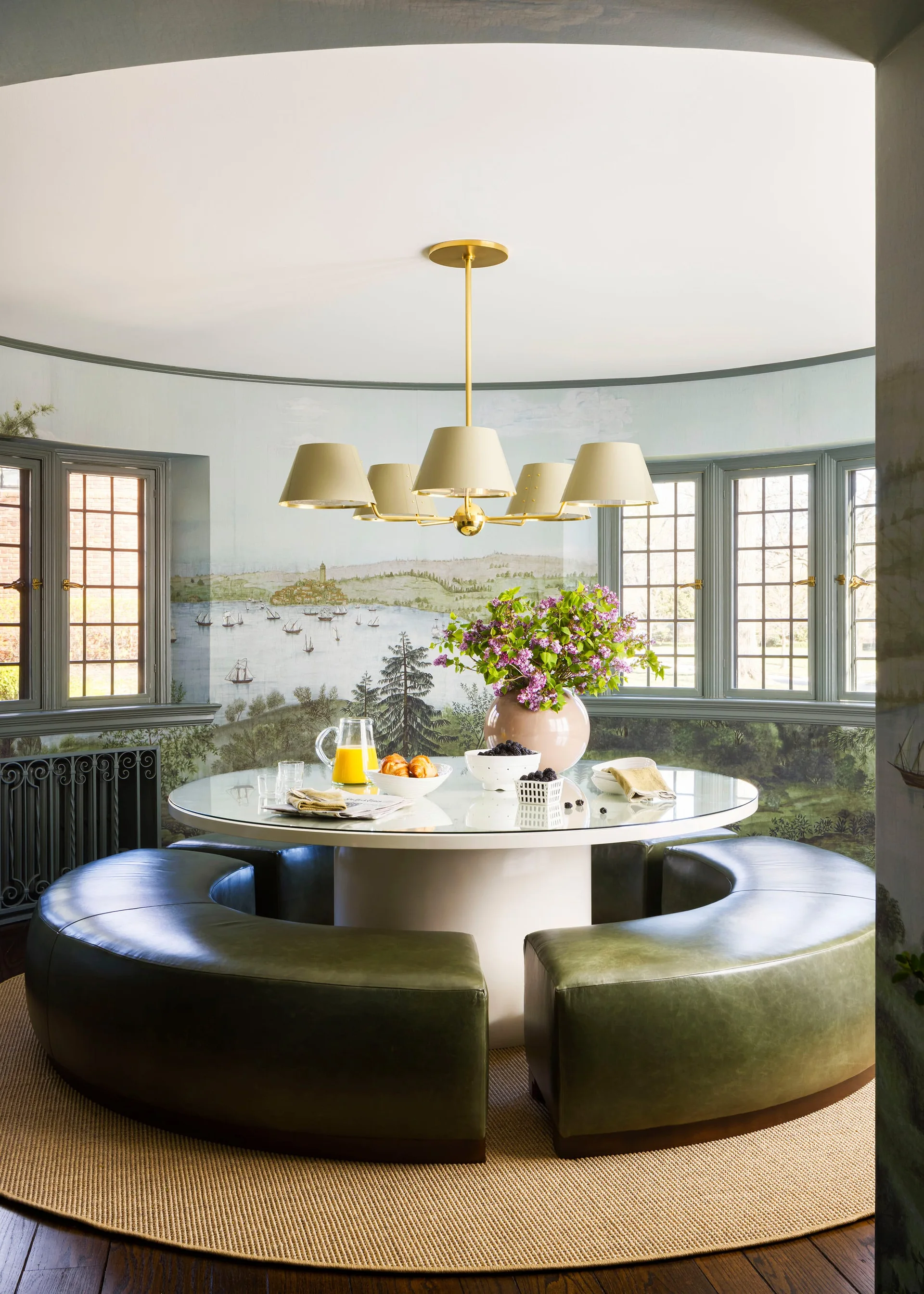 A circular glass-topped dining table with curved green leather banquette seating is set against a scenic landscape wallpaper mural.