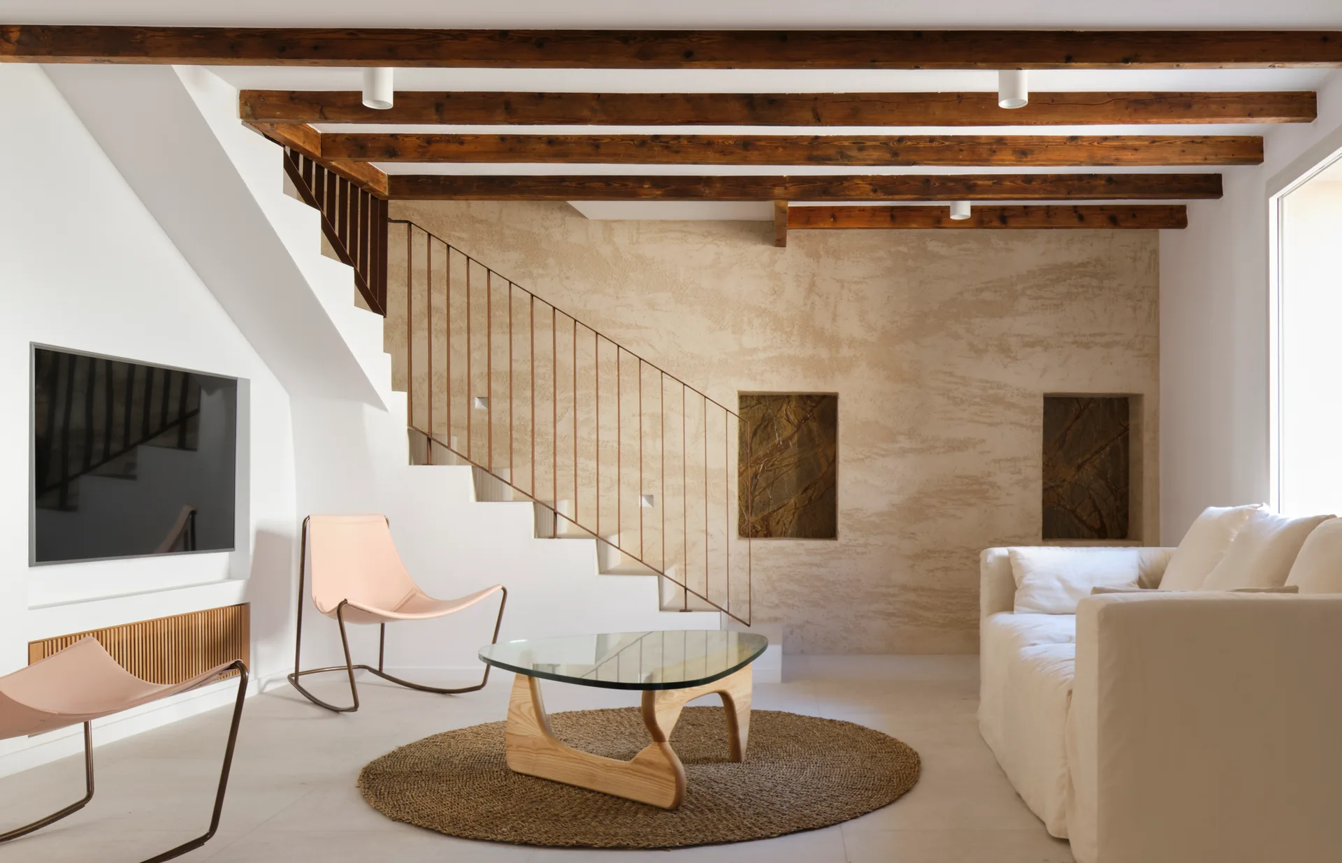 A minimalist living room features a white sofa, glass coffee table, two pink sling chairs, and a staircase against a textured plaster wall.