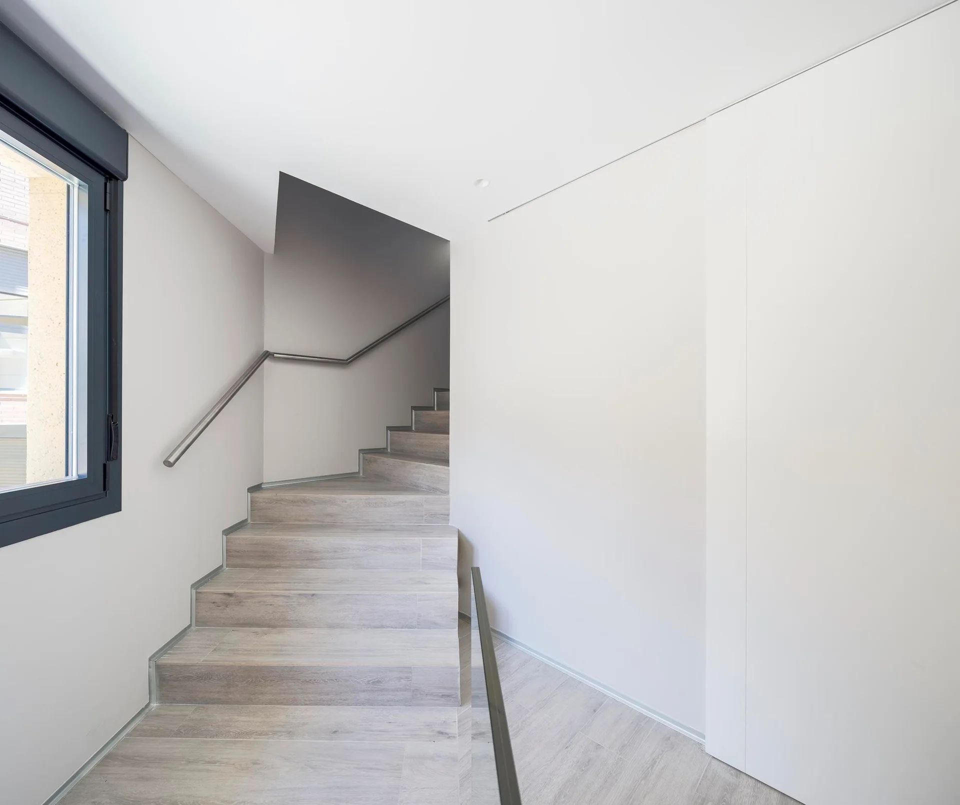 A minimalist staircase with light wood-grained treads and simple metal handrails ascends between bright white walls next to a window.