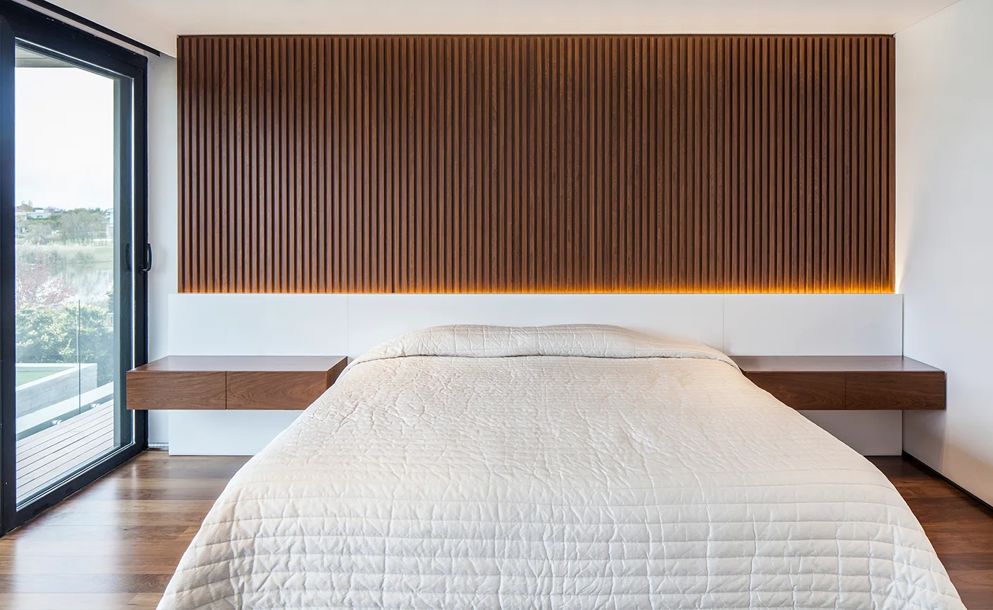 A minimalist bedroom featuring a vertical slatted wood accent wall, built-in white headboard, floating wood nightstands, and a quilted bed.