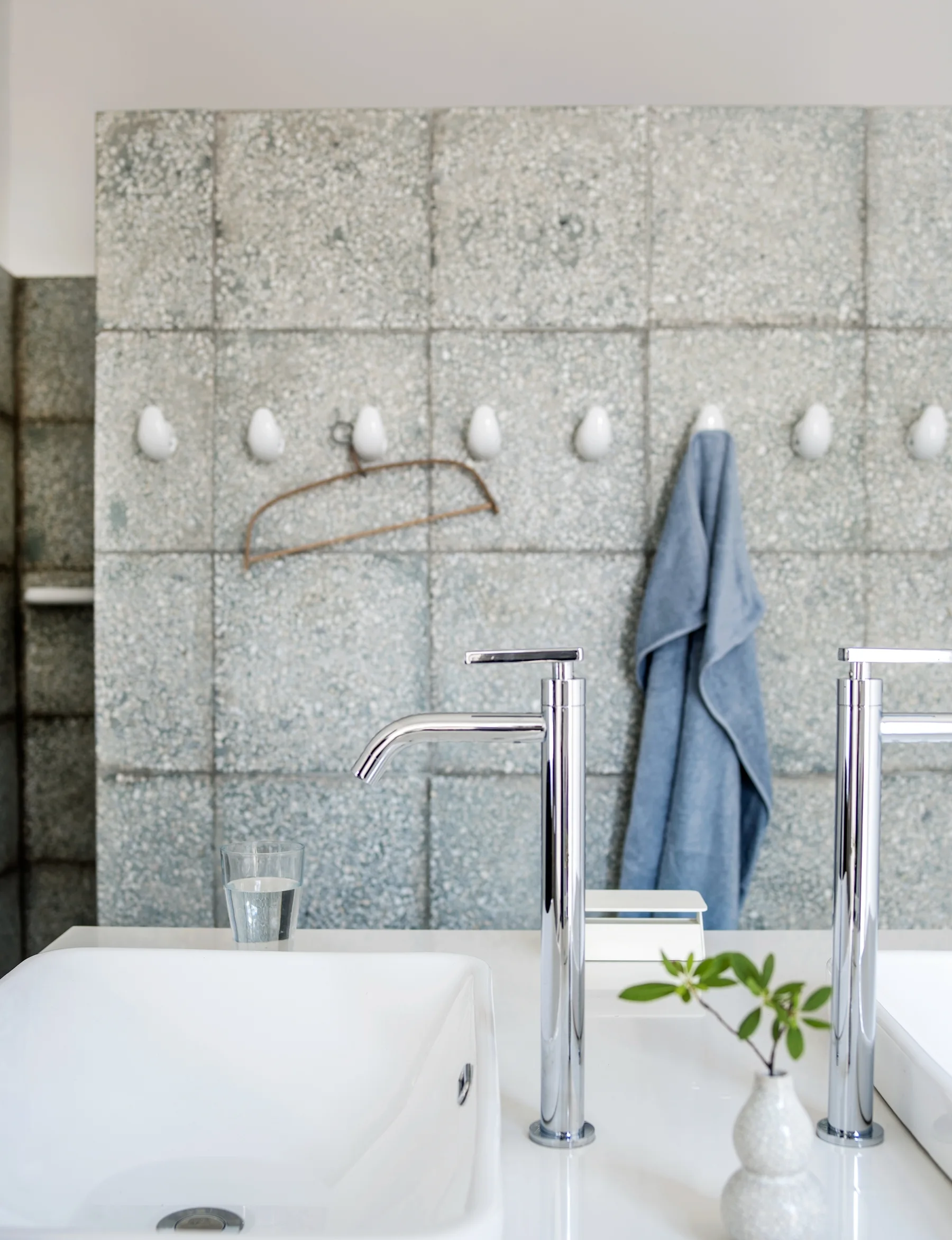 A bathroom vanity features two white rectangular sinks and tall chrome faucets in front of a grey speckled tile wall with a row of white hooks.