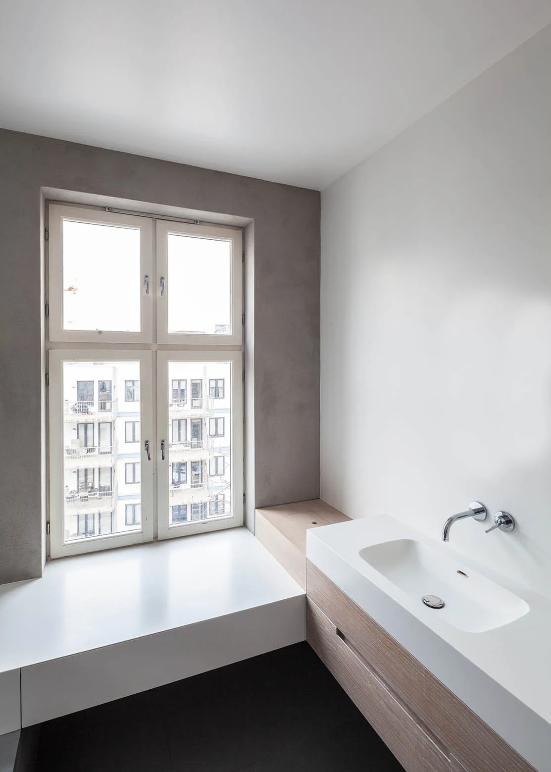 A minimalist bathroom features a wood vanity with an integrated white sink, a chrome wall faucet, and a large window in a grey plaster wall.