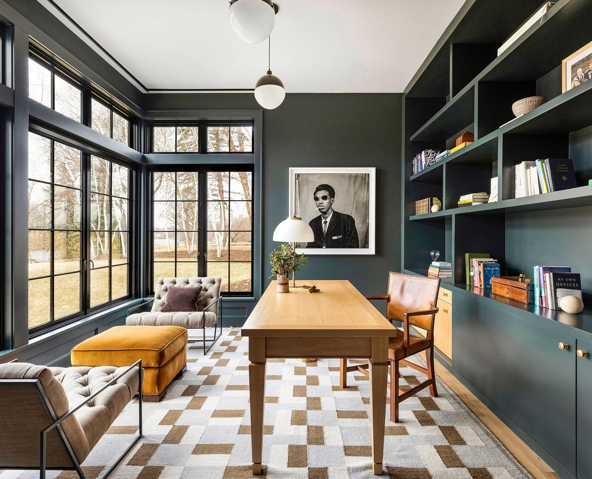A home office features a wood desk, a leather chair, dark green built-in bookshelves, and large grid windows looking out to trees.