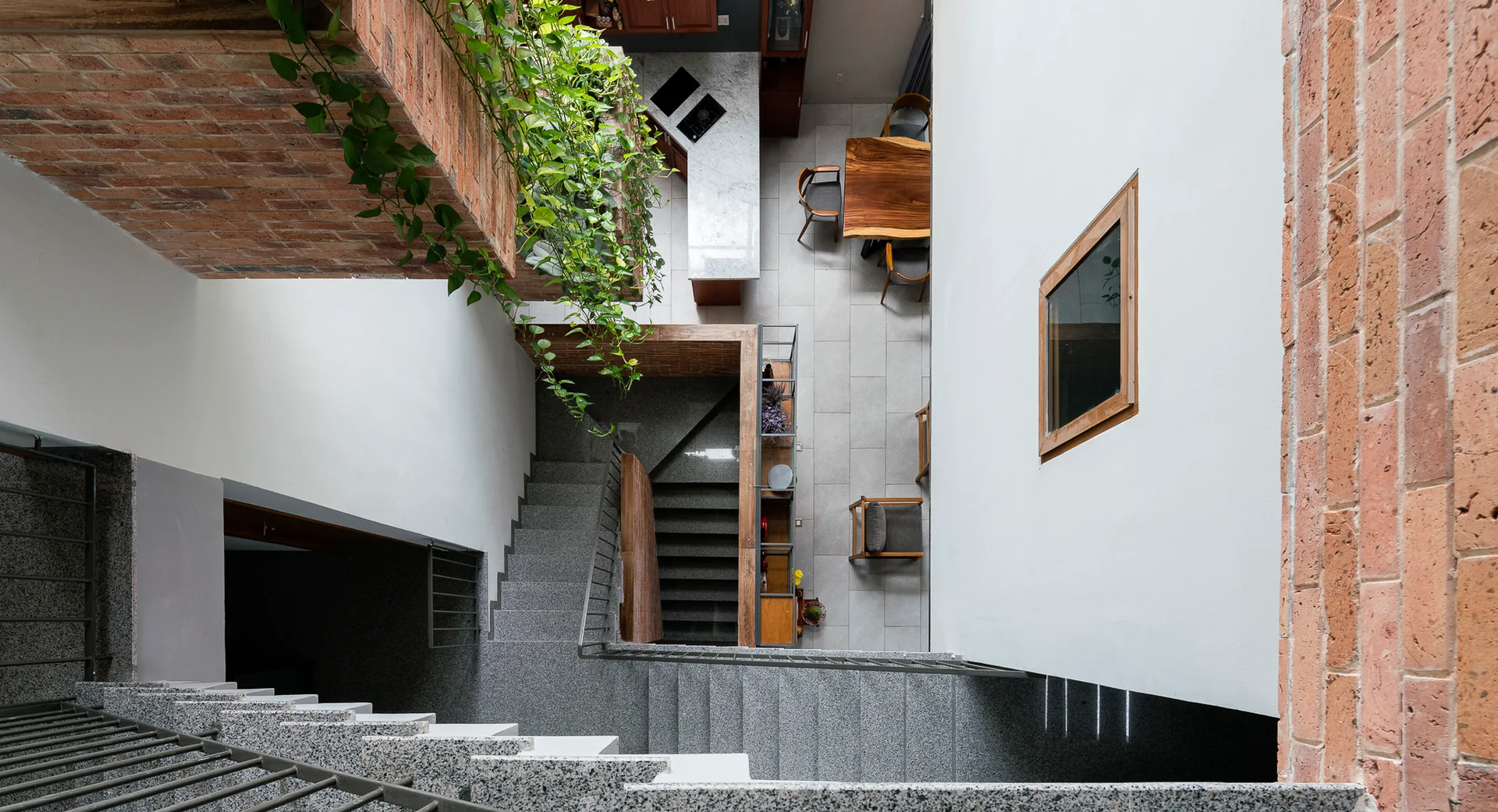 An overhead view of a grey stone staircase flanked by white and exposed brick walls, with green vines hanging from above overlooking a dining area.