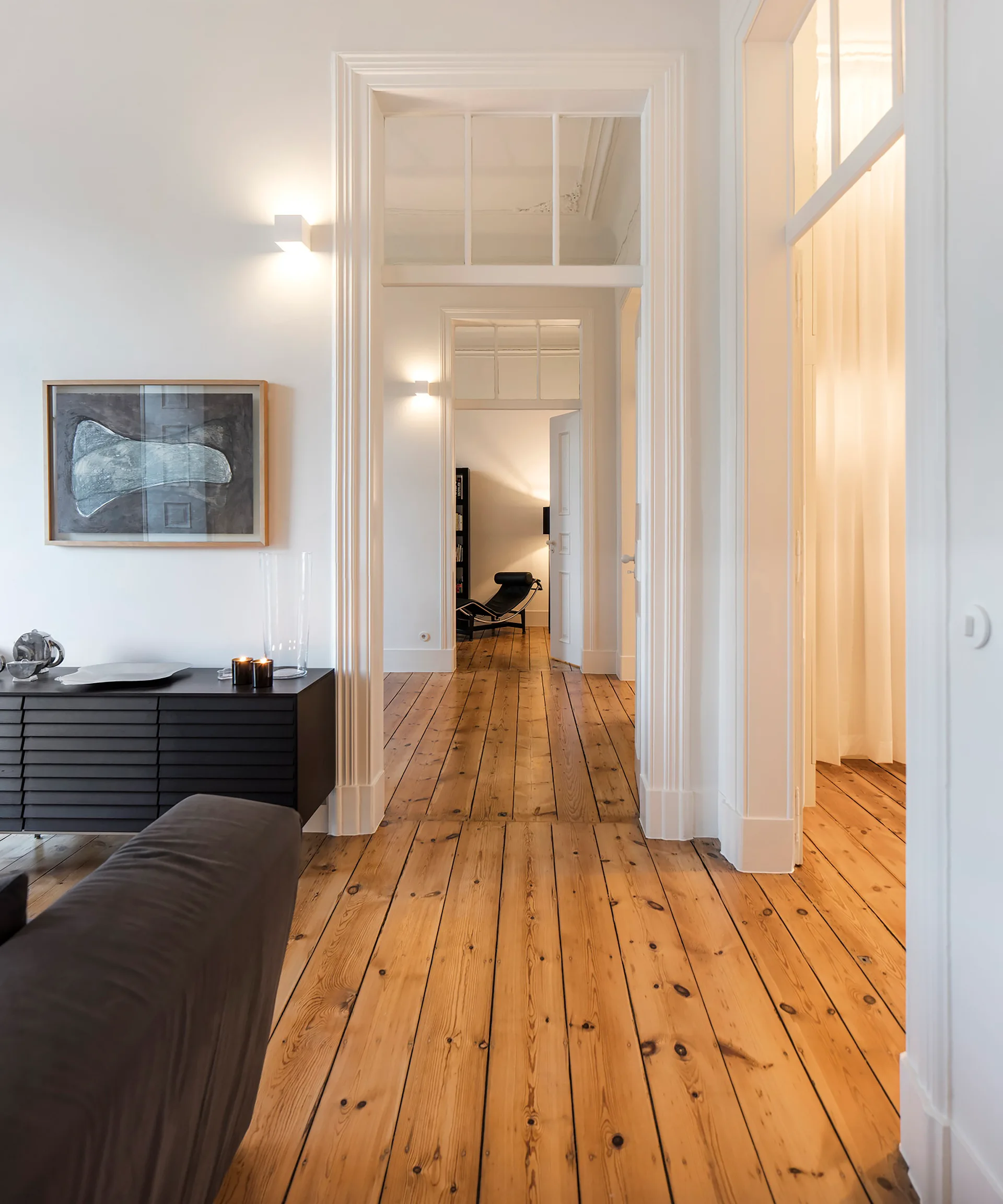 A view down a white apartment hallway with natural wood plank floors, tall doorways featuring transom windows, and a black sideboard.
