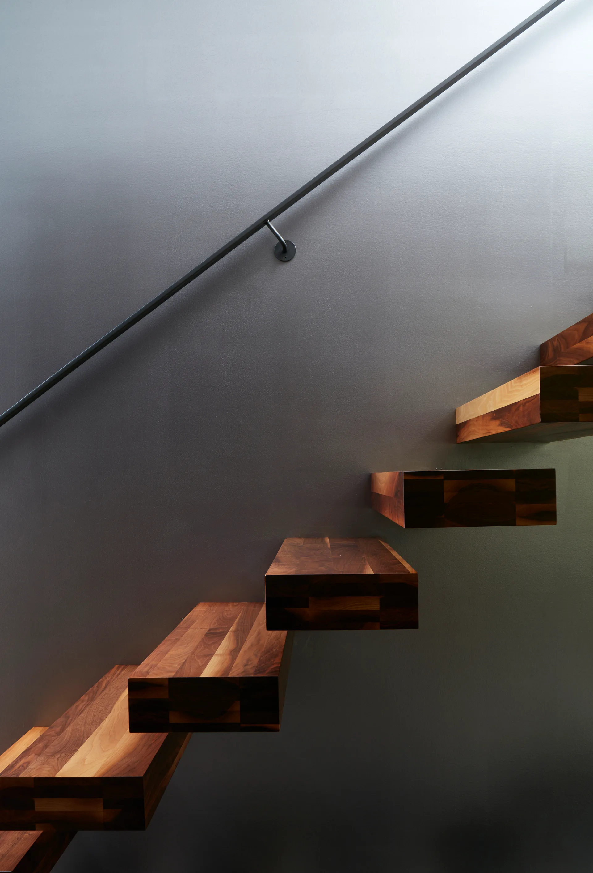 A minimalist floating wooden staircase with thick rectangular treads is mounted on a smooth gray wall next to a slim black handrail.