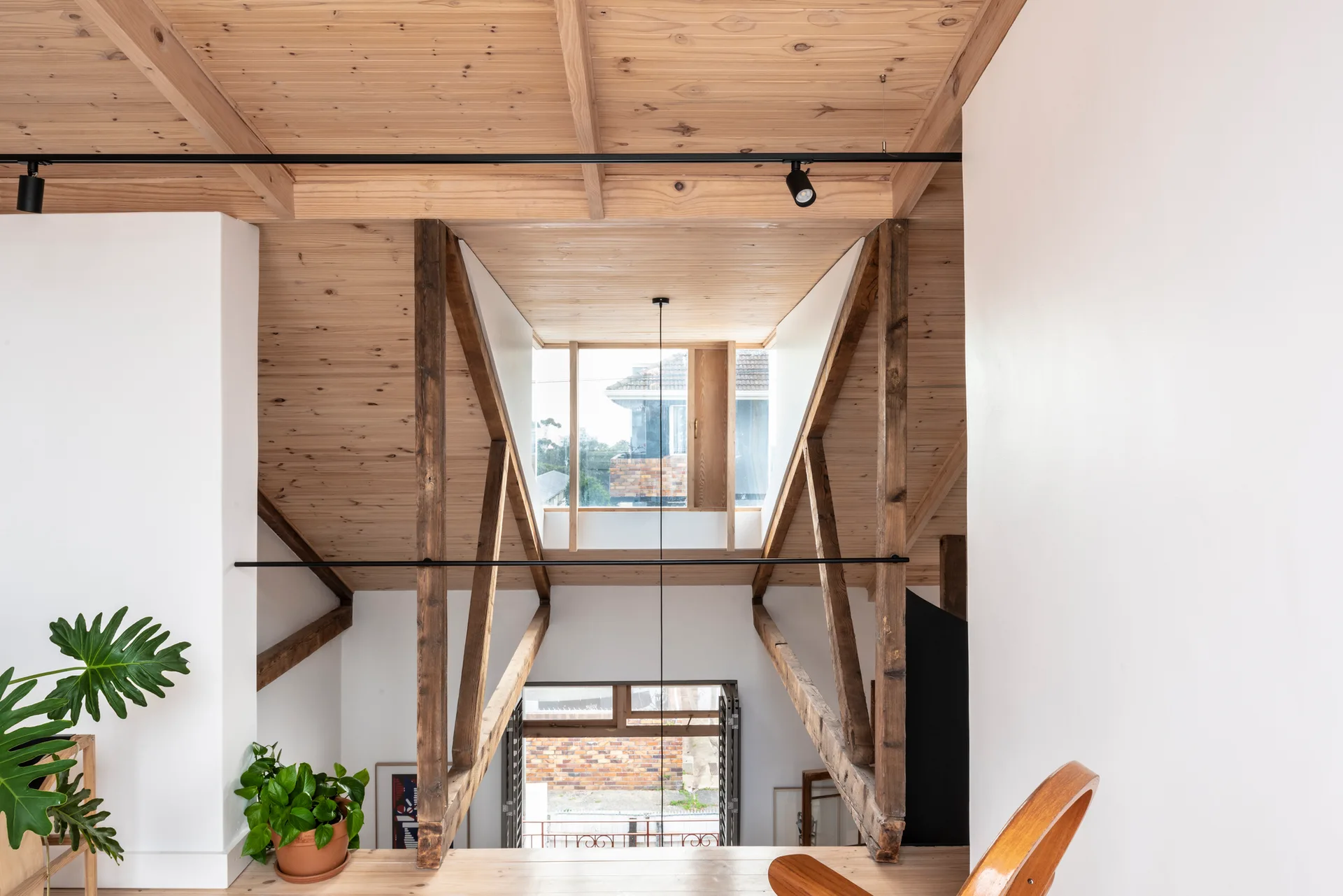 A loft interior features a vaulted light wood ceiling, dark timber structural trusses, white plaster walls, and black track lighting.