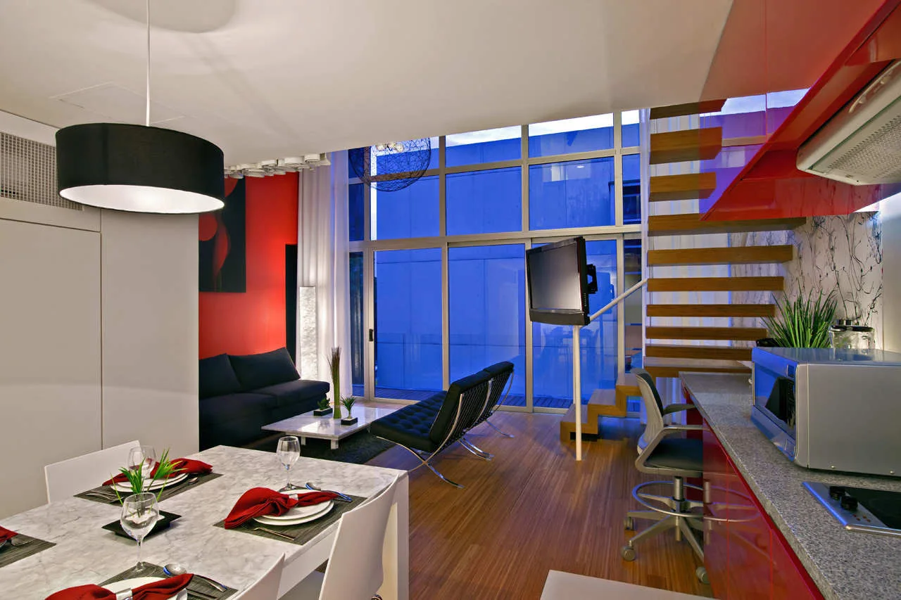 A modern open-concept apartment featuring a marble dining table, black sofa, floating wooden stairs, and glossy red kitchen cabinets.