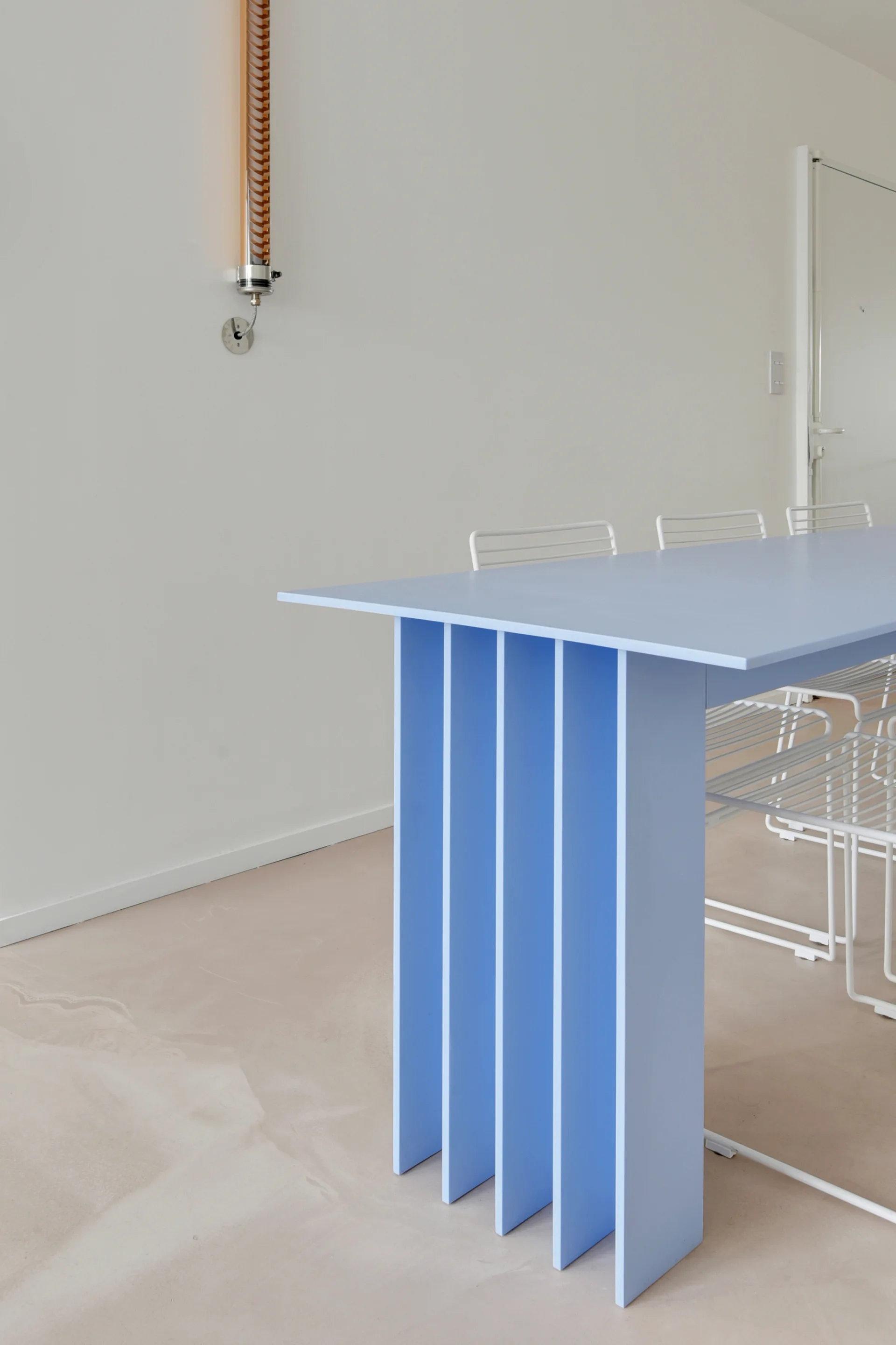 A light blue dining table with a slatted base is paired with white wire chairs against a plain white wall with a tubular wall sconce.