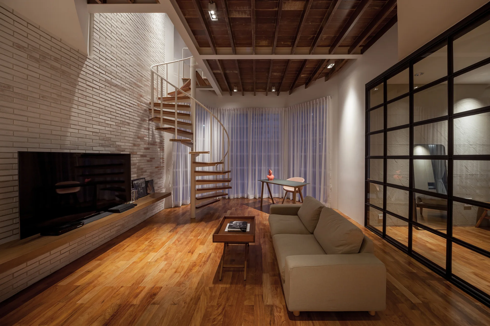 A modern living room features a beige sofa, a white metal spiral staircase, a white brick wall, and a black-framed glass partition.