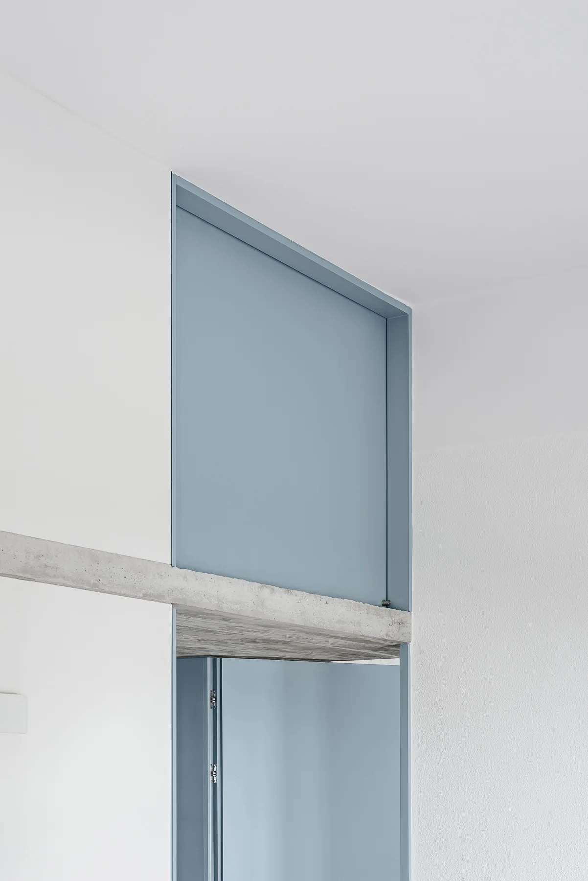 A minimalist white wall features a blue metal doorway frame integrated with a rough, exposed concrete lintel beneath a smooth white ceiling.
