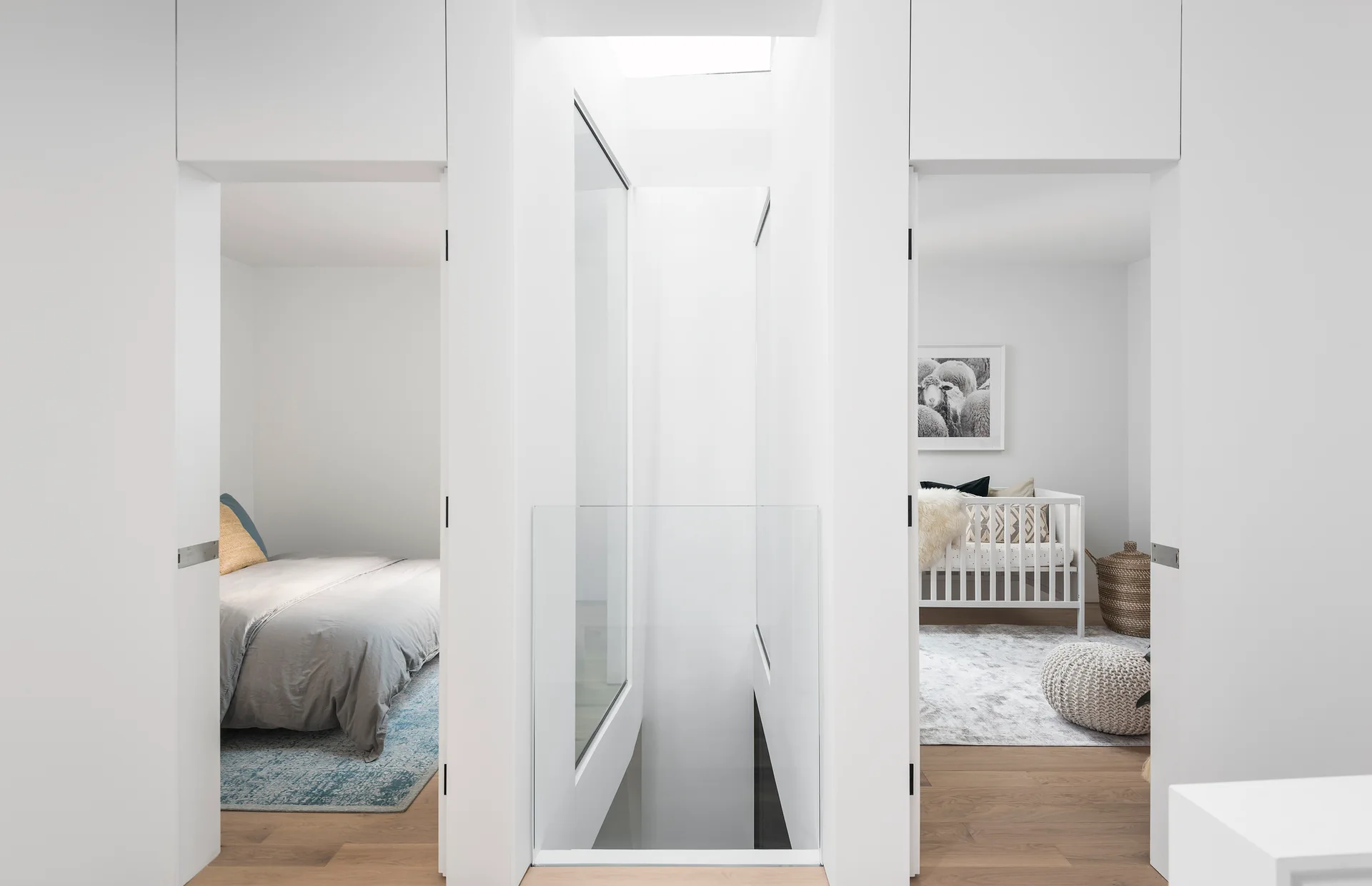A minimalist white landing with a glass staircase railing looks into a bedroom and a nursery with light wood flooring.