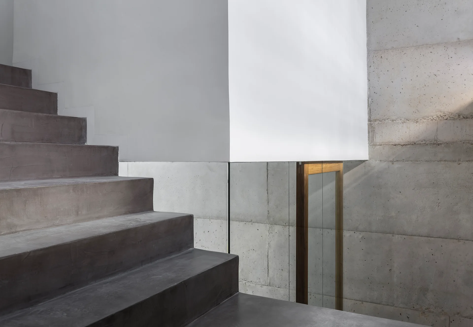 A minimalist concrete staircase rises next to an exposed concrete wall with a frameless glass railing.