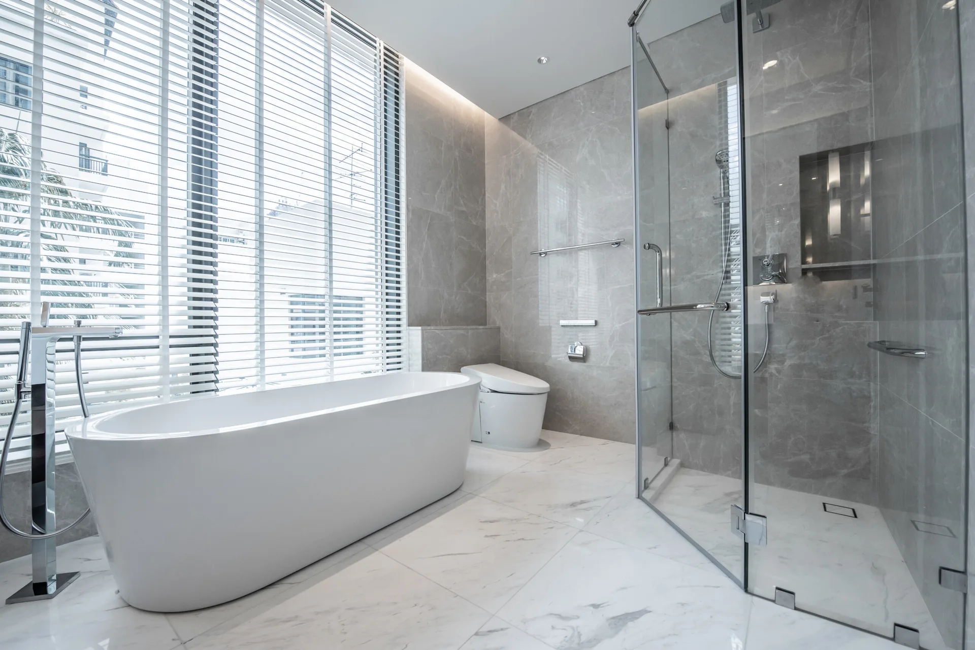A modern bathroom with a white freestanding bathtub by a window with blinds, a glass walk-in shower, and grey marble-patterned wall tiles.