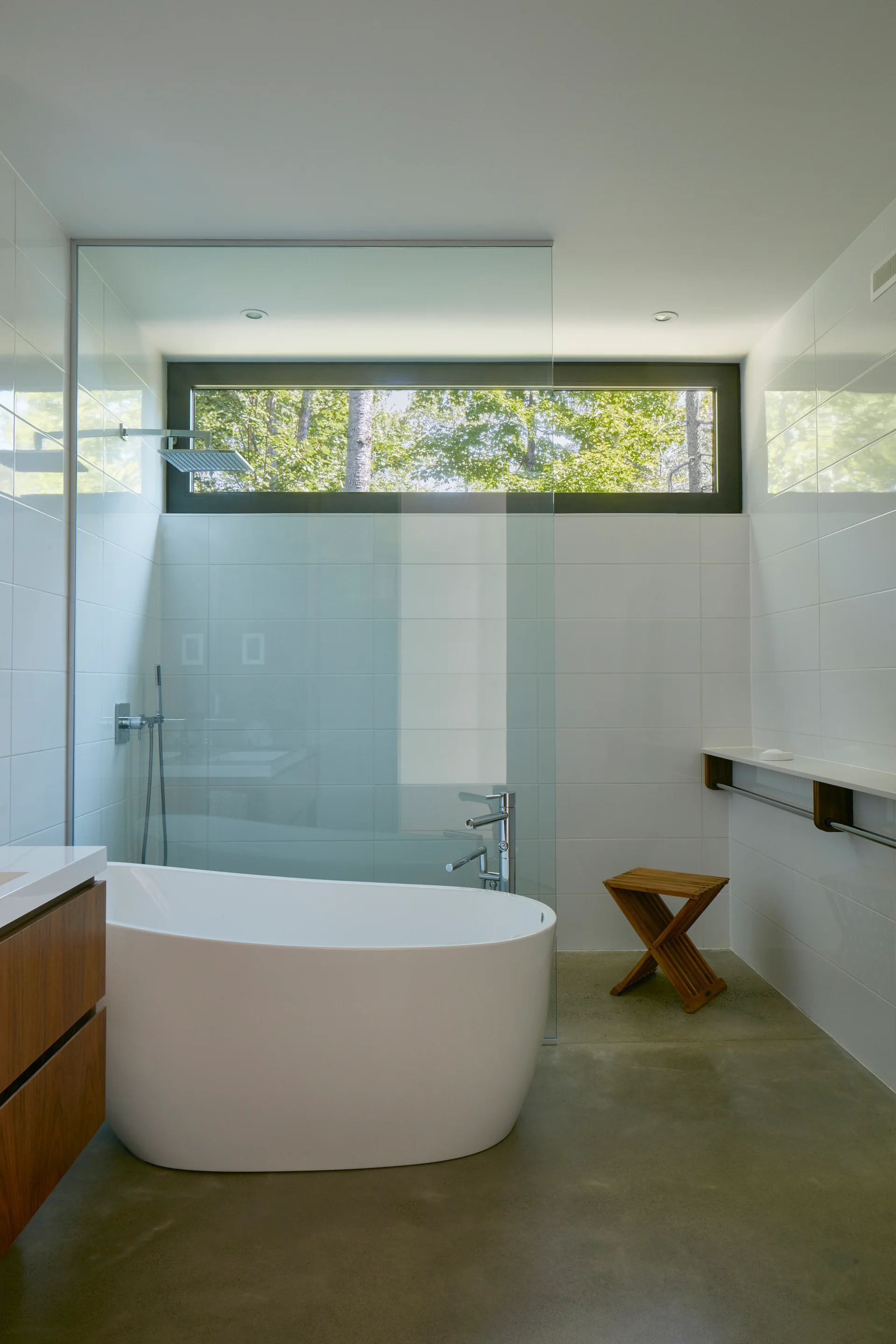A modern bathroom features a freestanding white bathtub, a glass shower partition, a concrete floor, and a horizontal window showing outdoor trees.