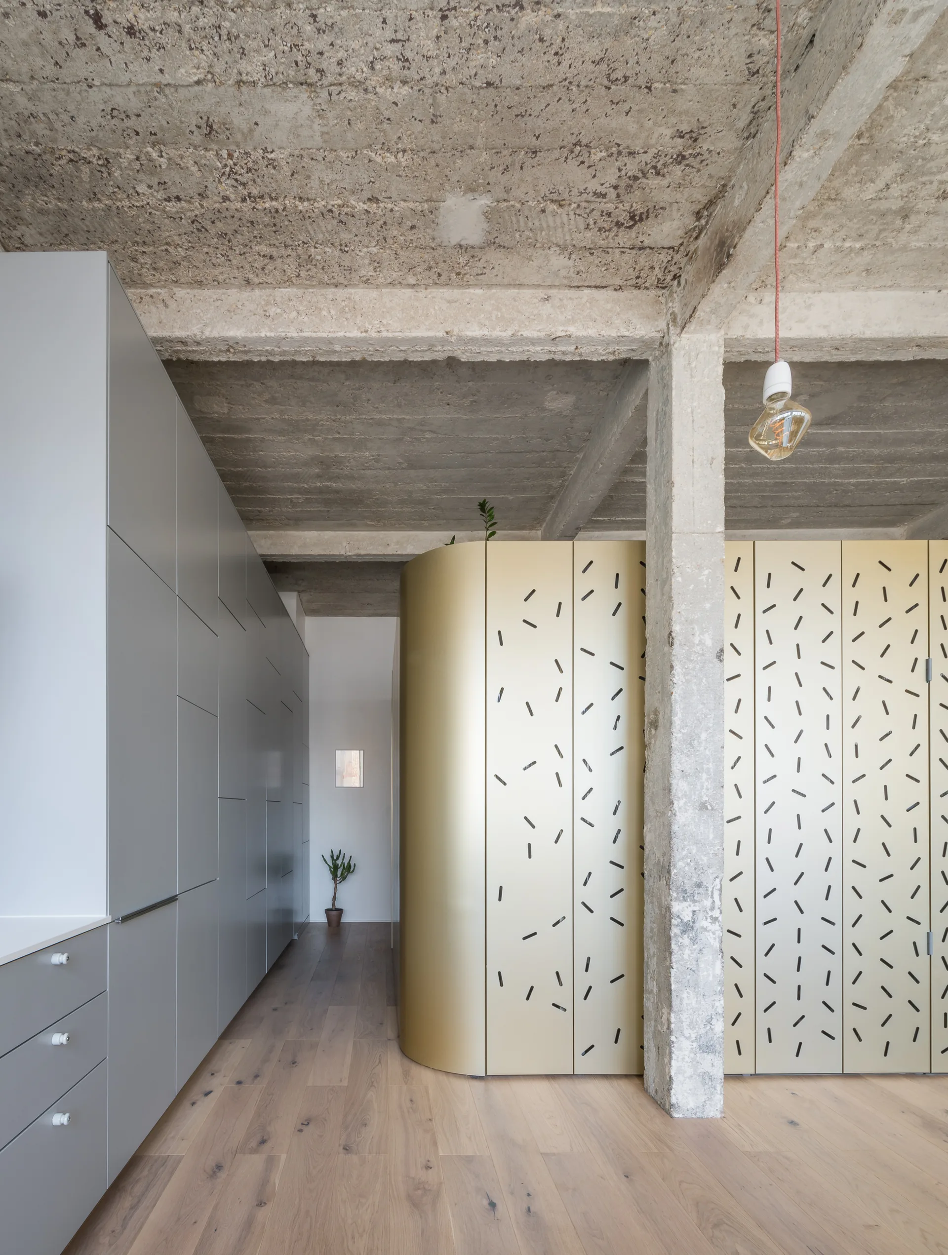 An interior space features light wood flooring, a raw concrete ceiling and column, grey built-in cabinets, and a curved metallic gold partition.