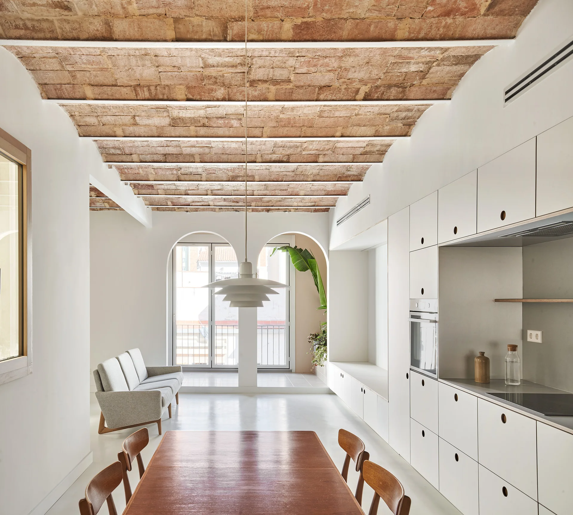 A minimalist kitchen and dining space featuring a vaulted brick ceiling, integrated white cabinetry, and a wooden dining table under a pendant light.