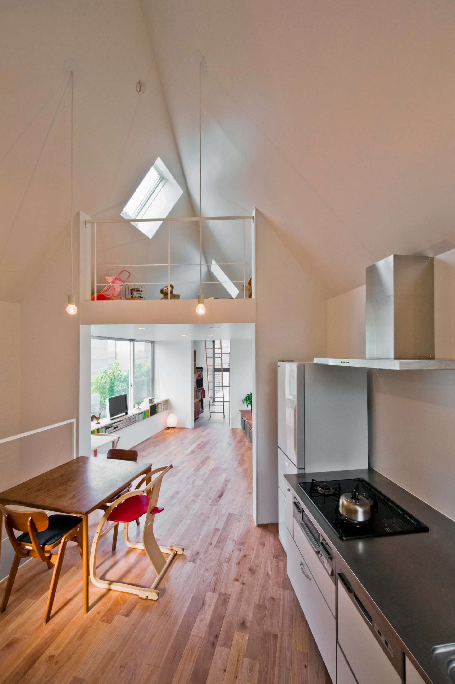 A minimalist white kitchen and dining area with hardwood floors, a vaulted ceiling with skylights, and a mezzanine loft above.
