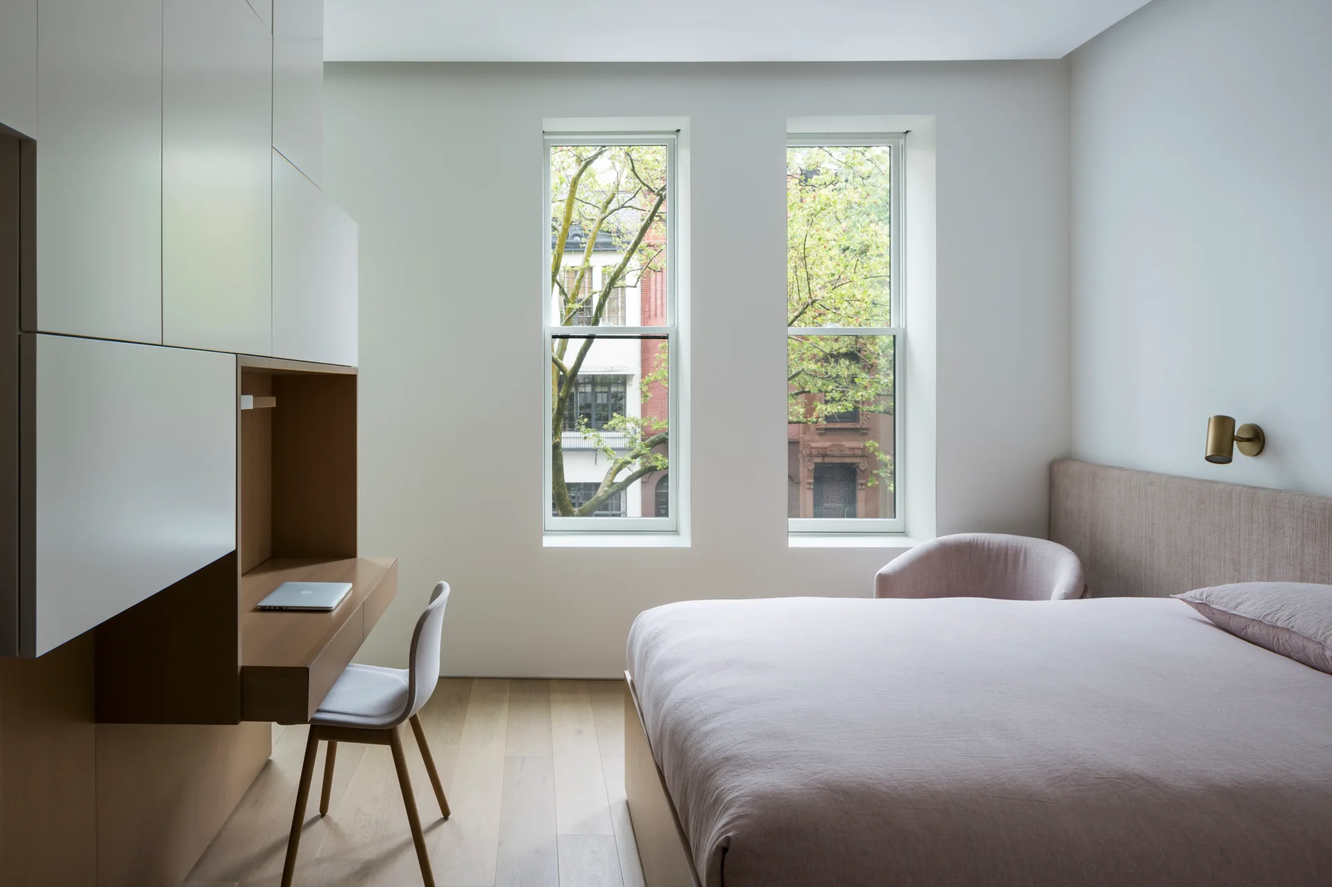 A modern minimalist bedroom features a custom built-in wood desk with white cabinetry, a bed with pale pink bedding, and two tall vertical windows.