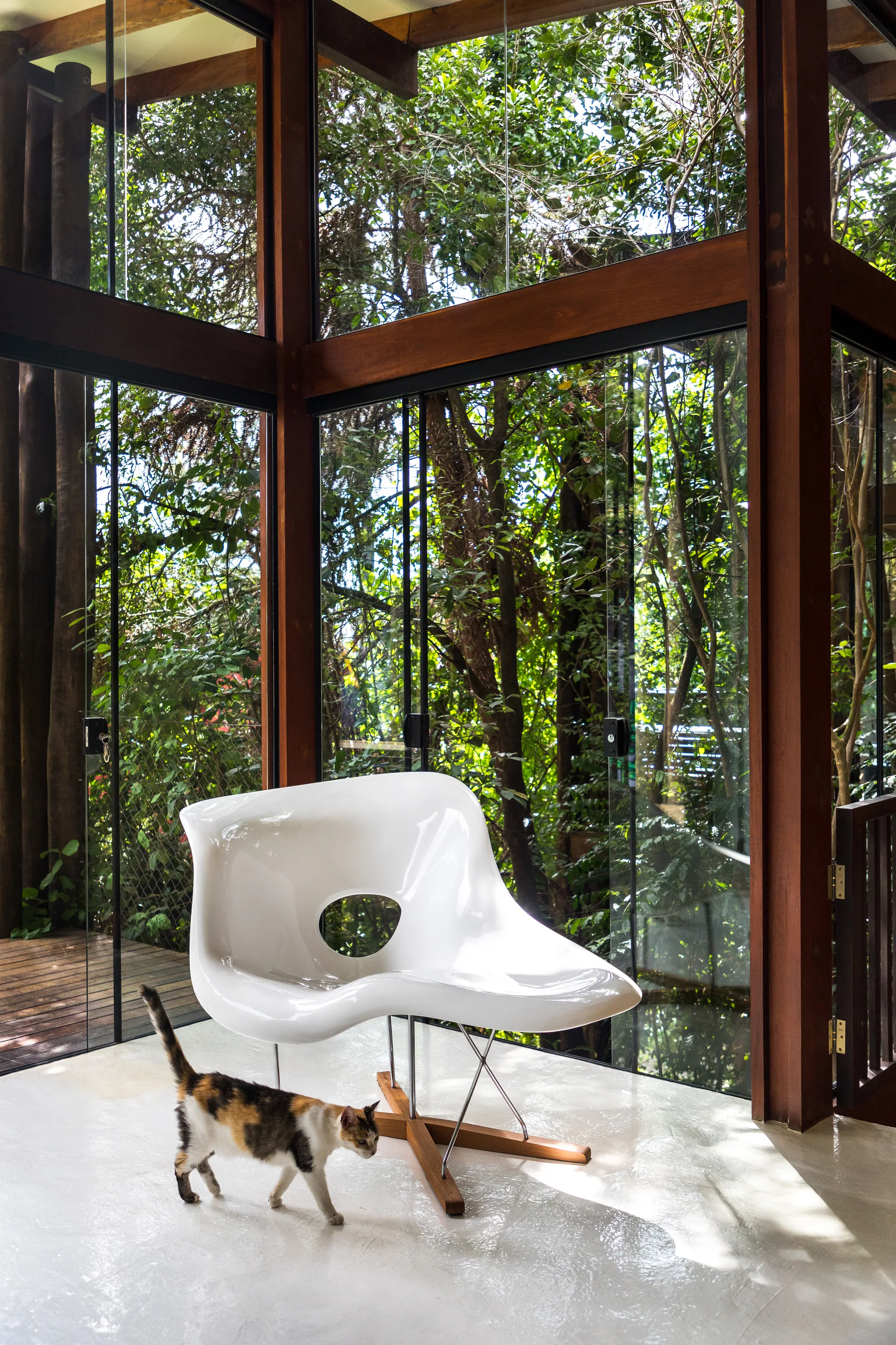 A sculptural white lounge chair sits on a glossy white floor in a room with floor-to-ceiling glass walls and wood frames.