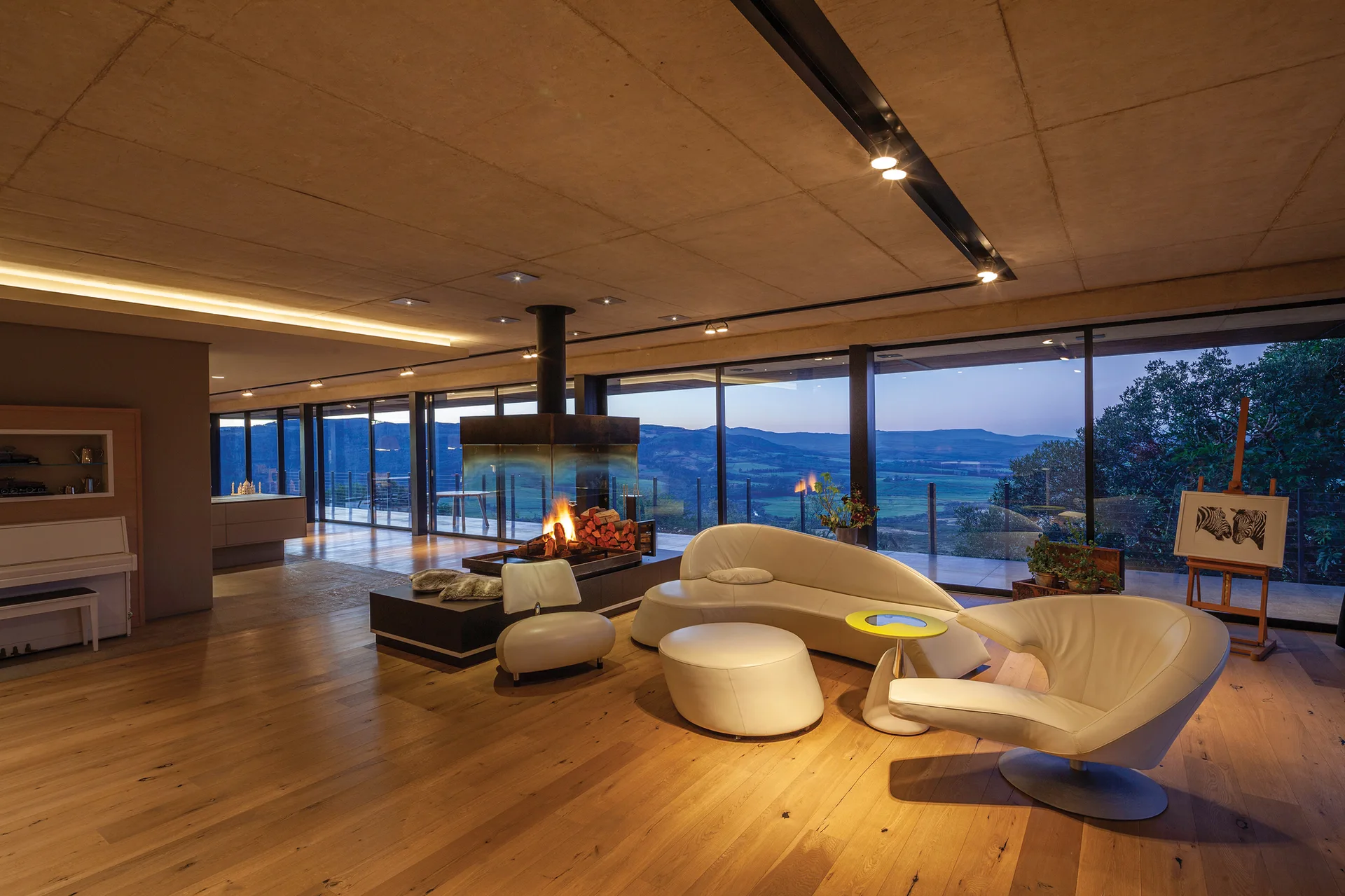 A spacious living room features curved white leather furniture, a central glass fireplace, and floor-to-ceiling windows showing a landscape at twilight.