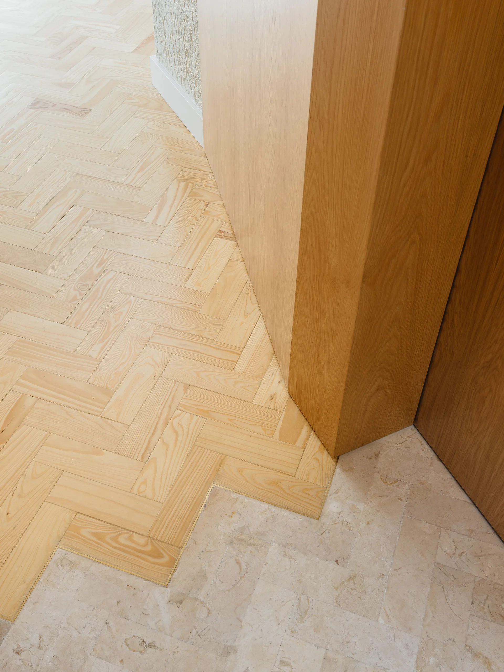 A close-up view of a floor transition where light wood herringbone parquet meets textured beige stone tiles laid in the same pattern.