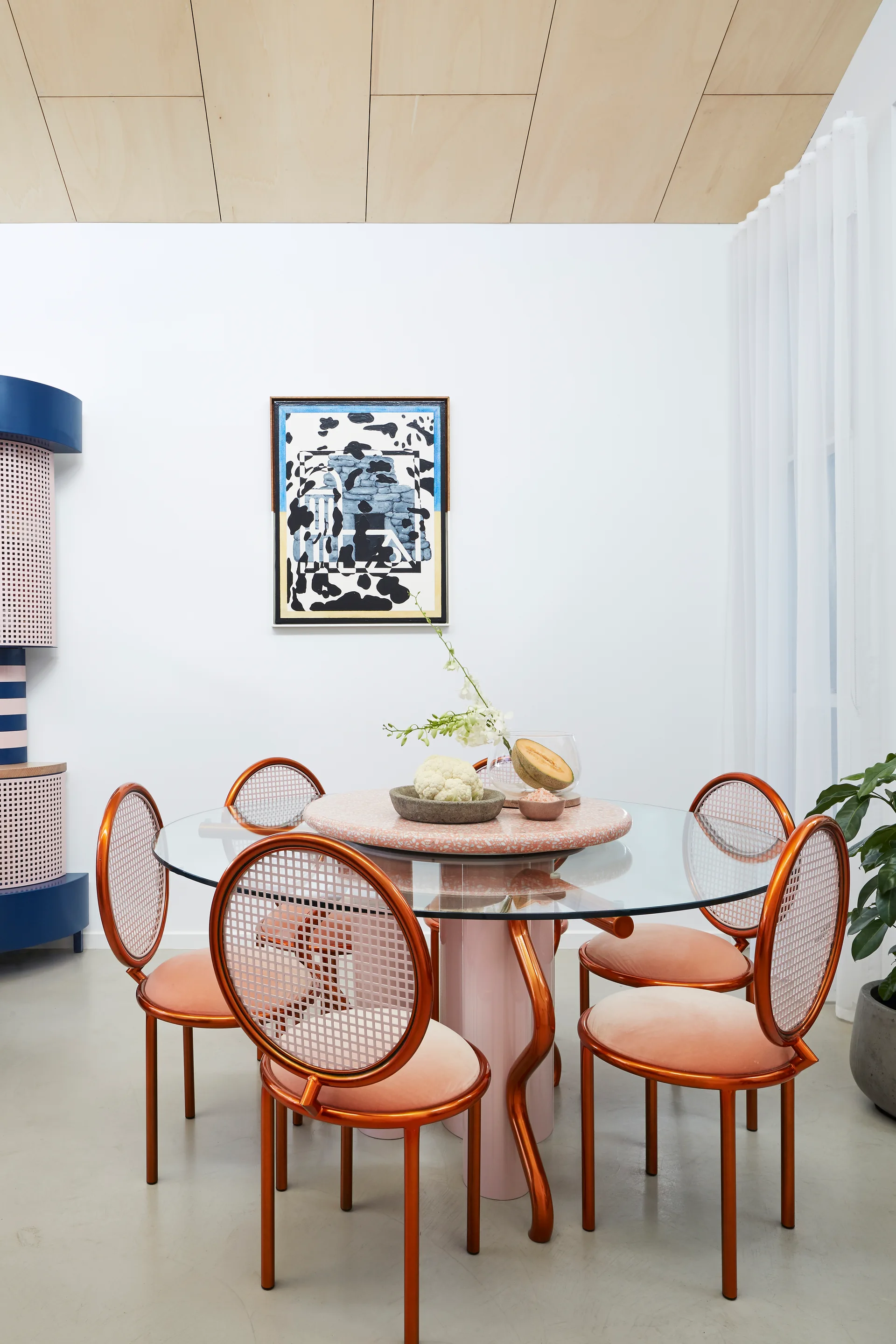 A glass-top dining table surrounded by orange metal cane-back chairs with pink velvet seats under a wood-paneled ceiling.