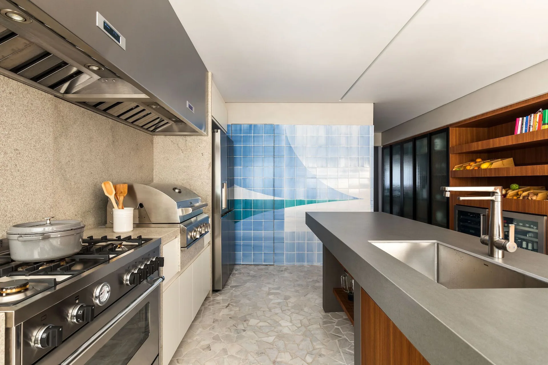 A modern kitchen features a grey island with a sink, a stainless steel stove, speckled stone flooring, and a blue geometric tile accent wall.
