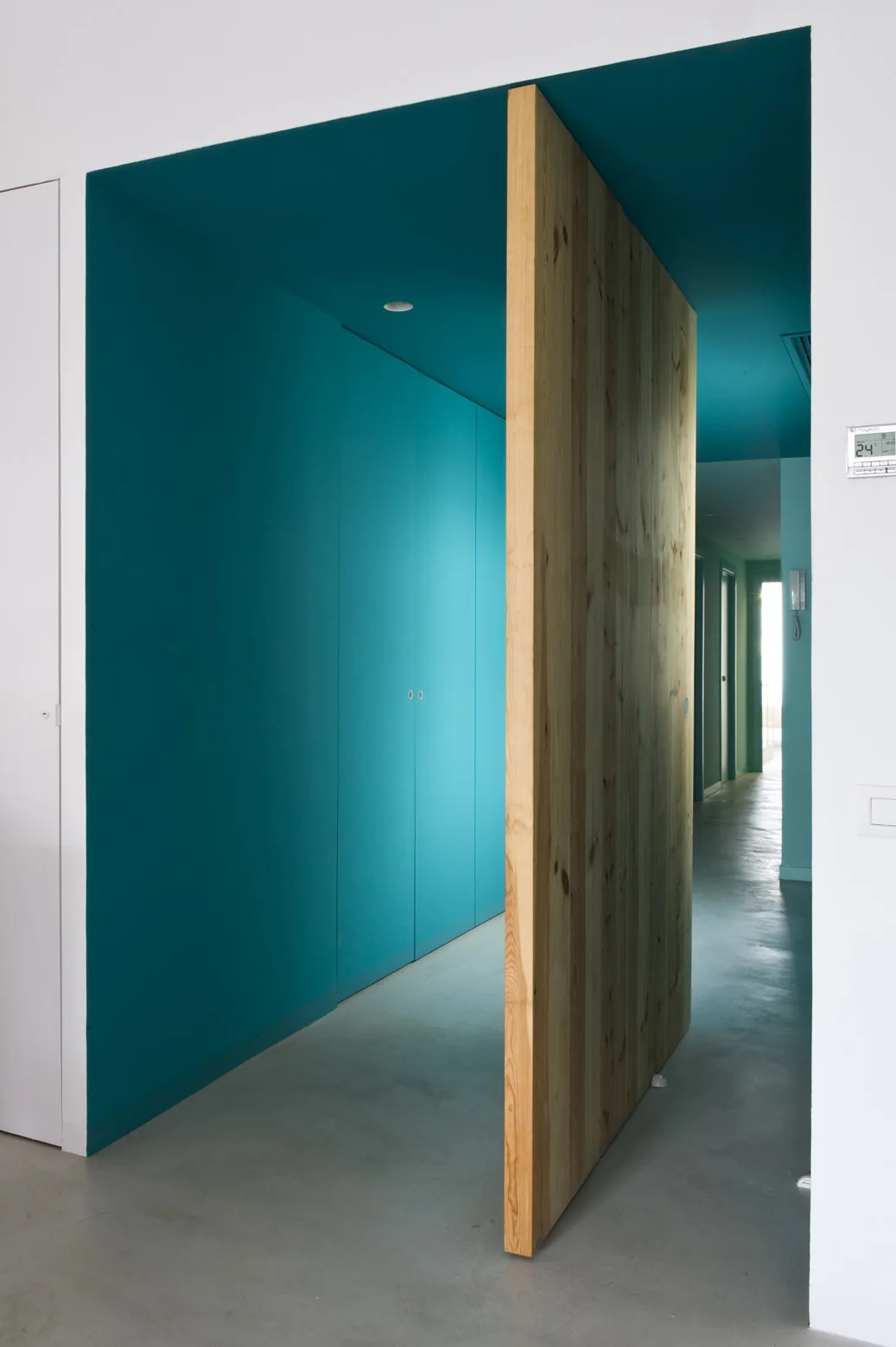 A minimalist hallway features a large, thick wooden pivot door integrated into vibrant teal painted walls and ceiling with a smooth grey concrete floor.