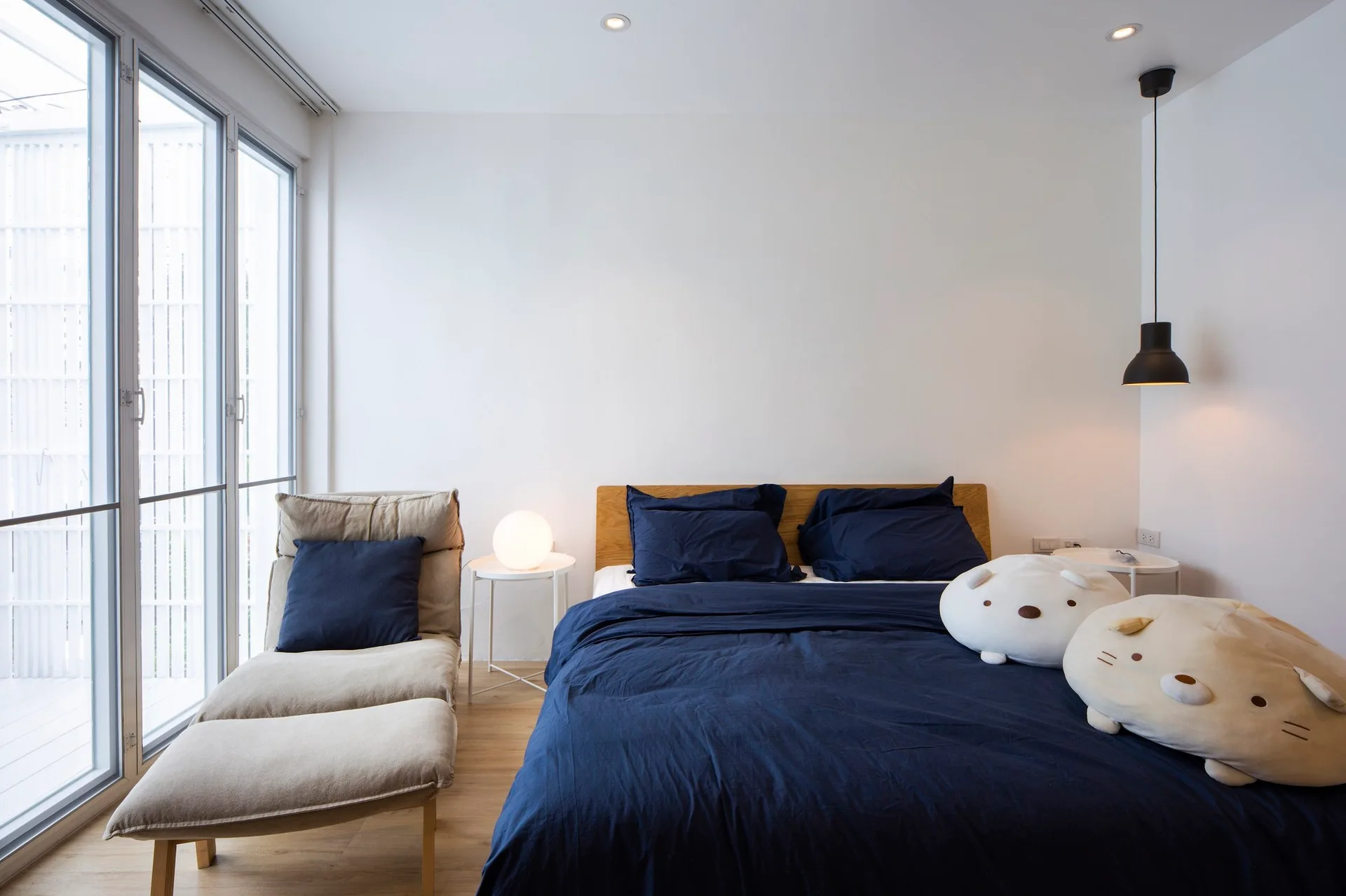 A minimalist bedroom features a bed with navy blue linens, two white side tables, and a beige lounge chair next to a large window.