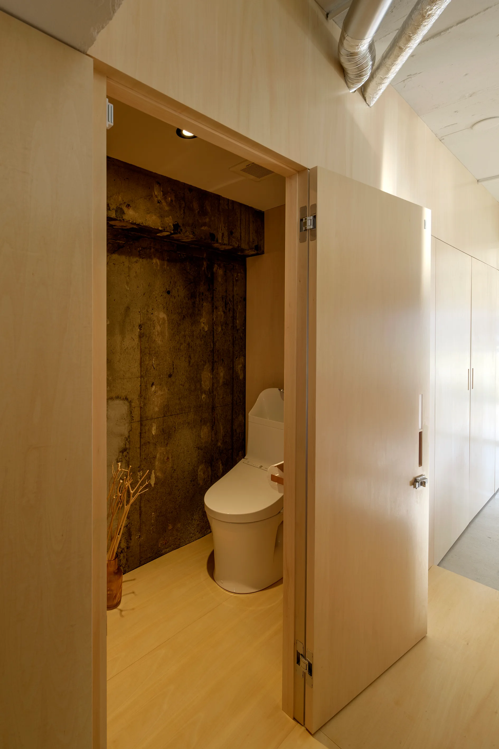 A minimalist powder room with light birch plywood walls, an open wooden door, a raw concrete back wall, and a white toilet.