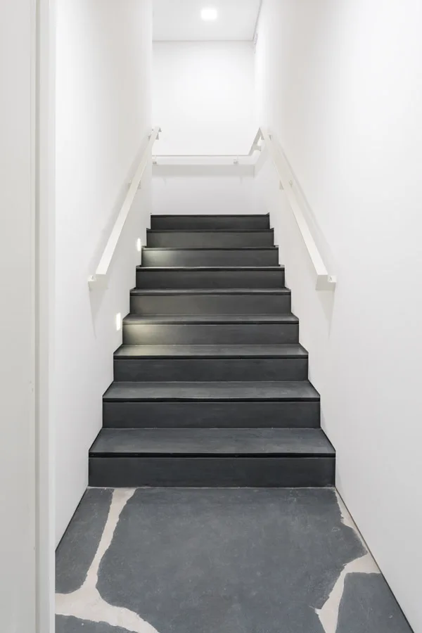 A minimalist staircase features dark matte treads, white walls, matching handrails, and small recessed step lights.
